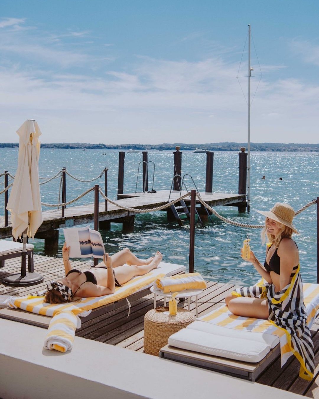 Lakeside living on Lake Garda Italy - two girls/women drinking cocktails overlooking the sparkling blue waters of the lake sitting on a pier belonging to AQVA Boutique Hotel Stylish Travel to Italy: Boutique Hotels, Design Stays & Culture