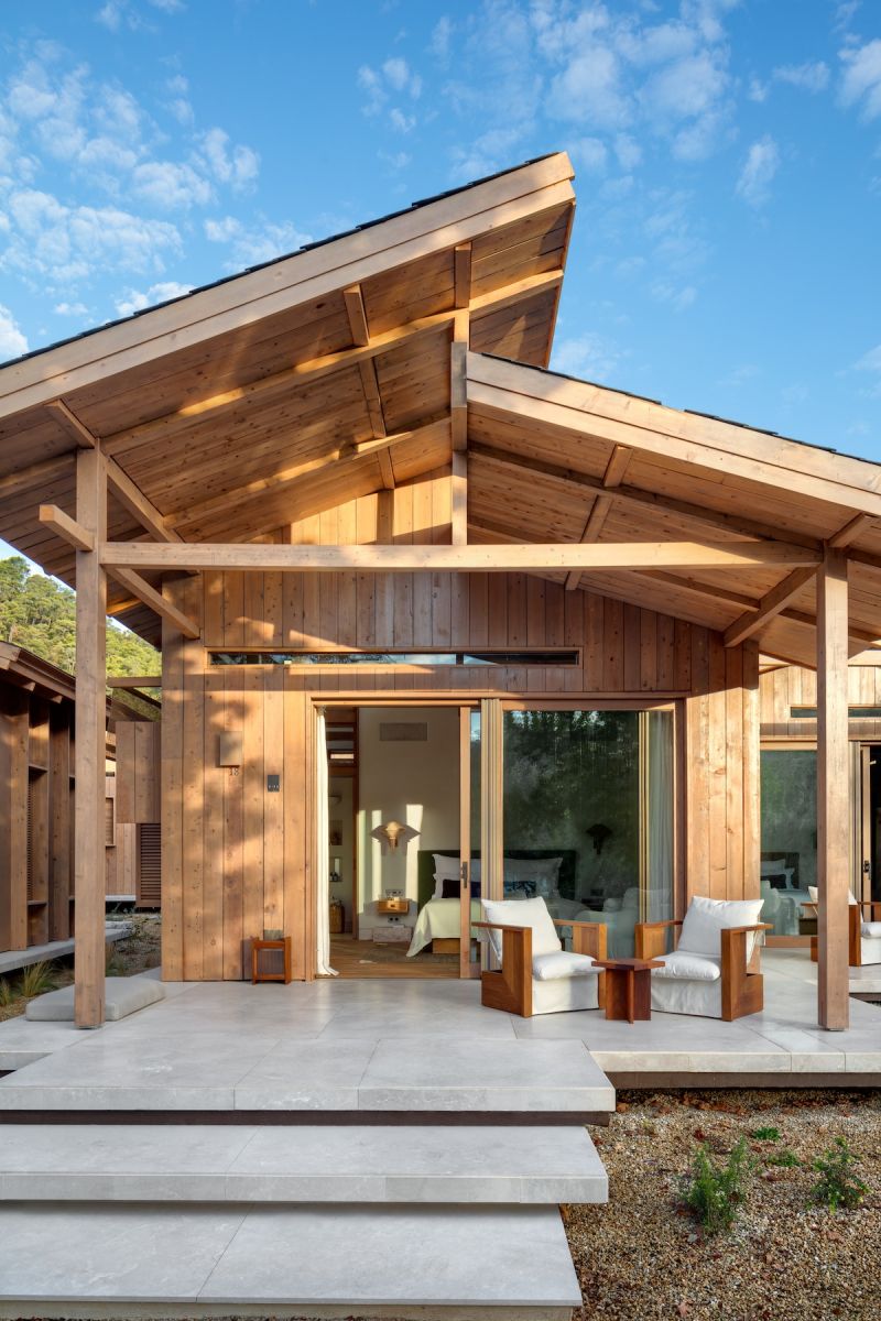 A pitched cedar-clad guest pavilion at AHÃMA, Muğla, its sliding glass wall opening to twin armchairs on a stone terrace under a timber-framed gable.