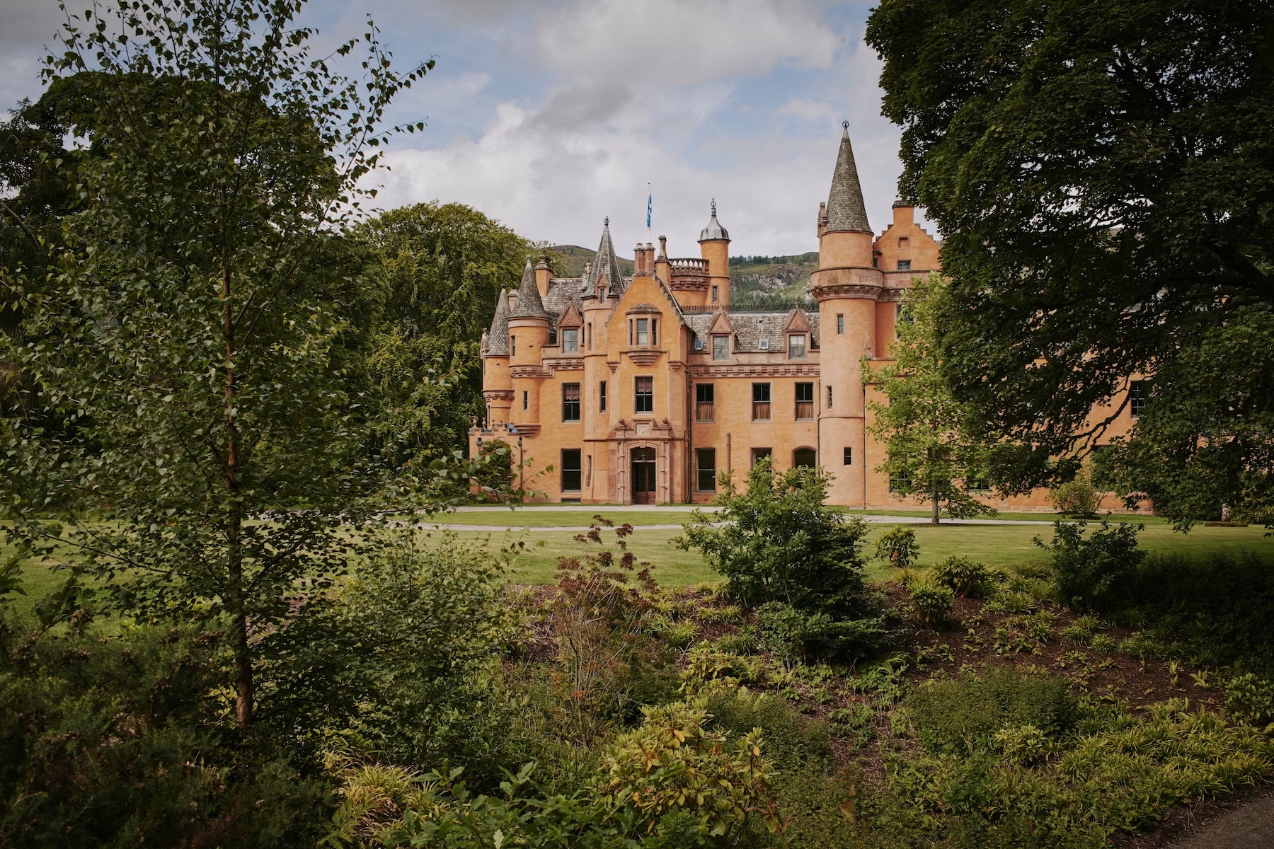 Aldourie Castle on the southern shore of Loch Ness, an apricot-pink Scottish baronial castle with turrets and Gothic detailing, framed by mature trees and gardens.