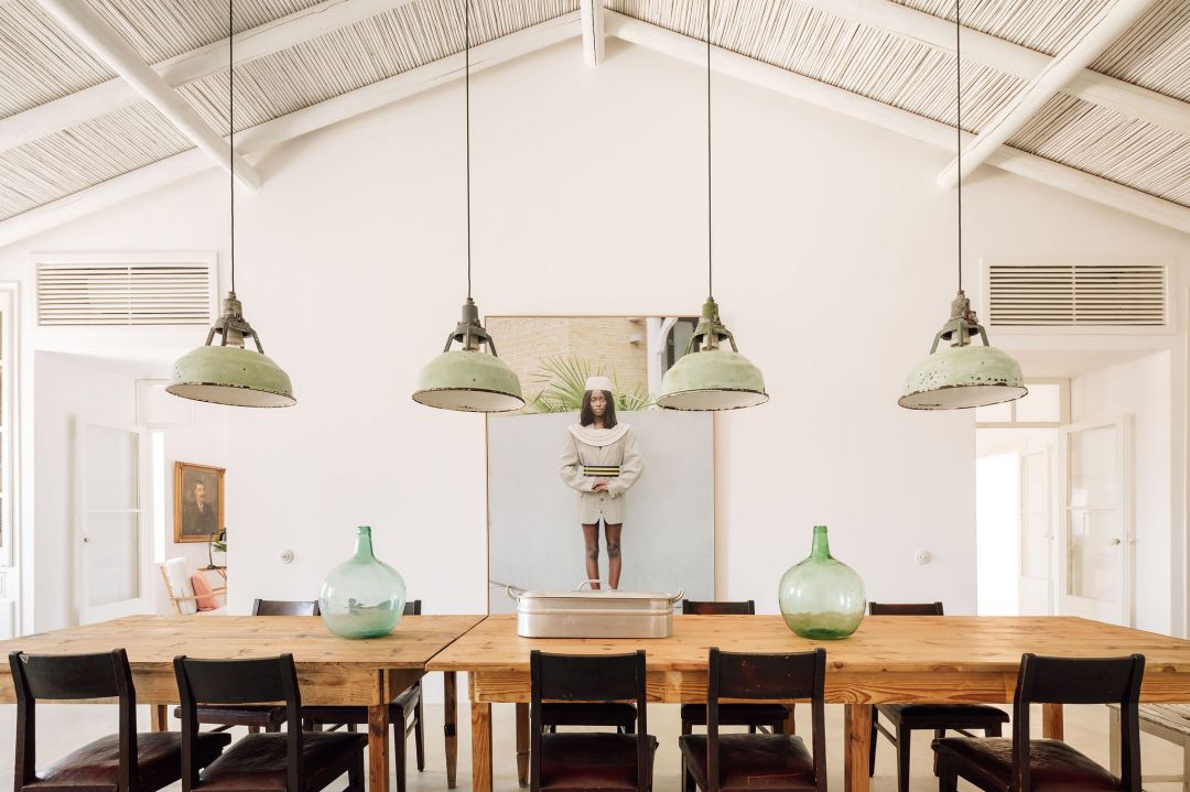 The communal dining room at Hospedaria in Tavira, with a long pine table and dark chairs beneath green industrial pendant lamps, a vaulted reed ceiling and green glass demijohns, a framed artwork on the far wall.