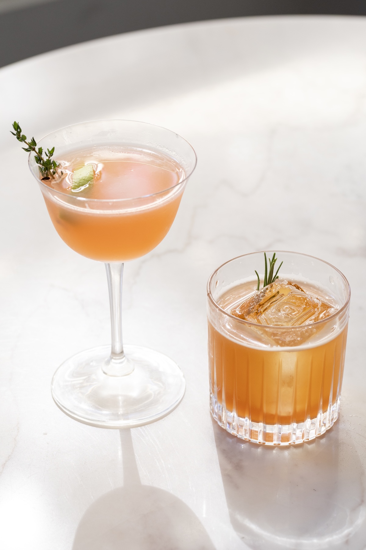 Two Pop-Up Peruano cocktails on a marble table, a coupe garnished with thyme and a rocks glass over a single large ice cube garnished with rosemary.