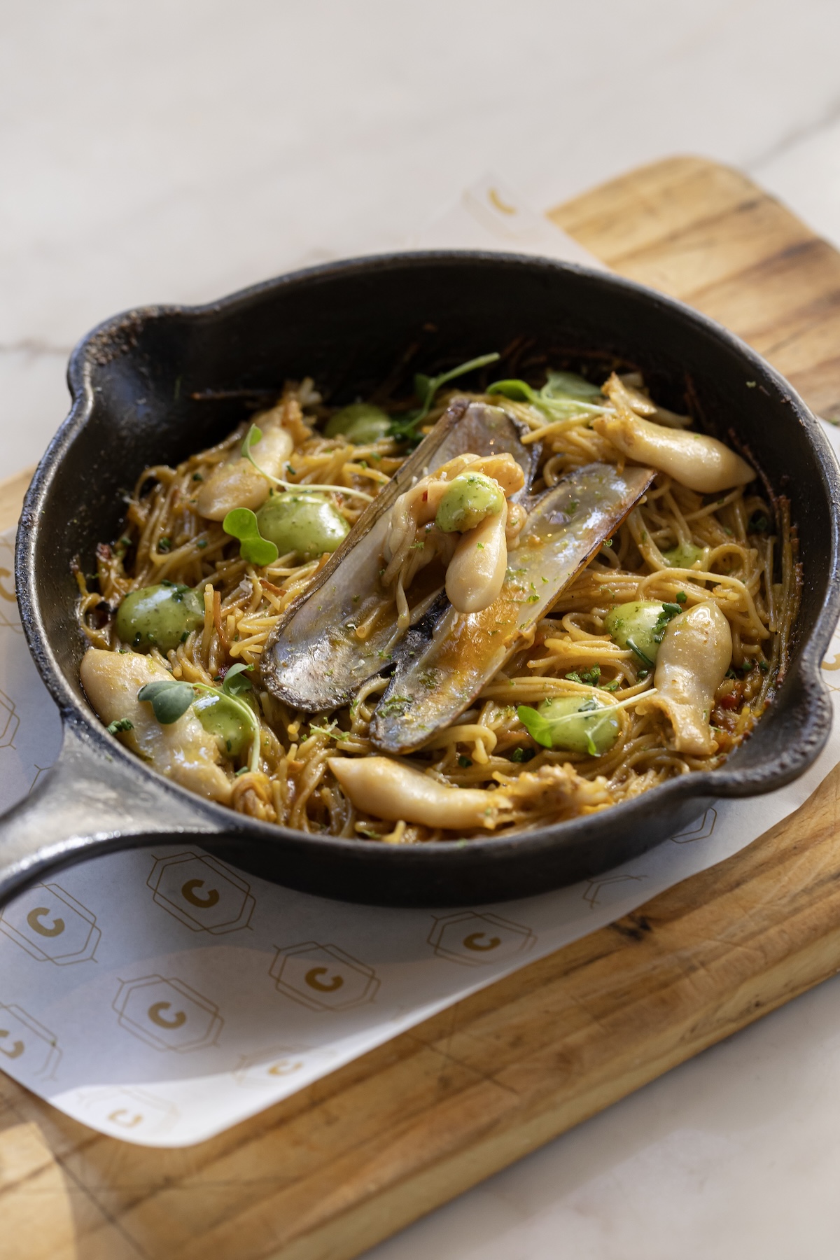 Ria Formosa seafood fideuá at Pop-Up Peruano, fine toasted noodles with a razor clam and herb oil, served in a cast iron pan on a wooden board.