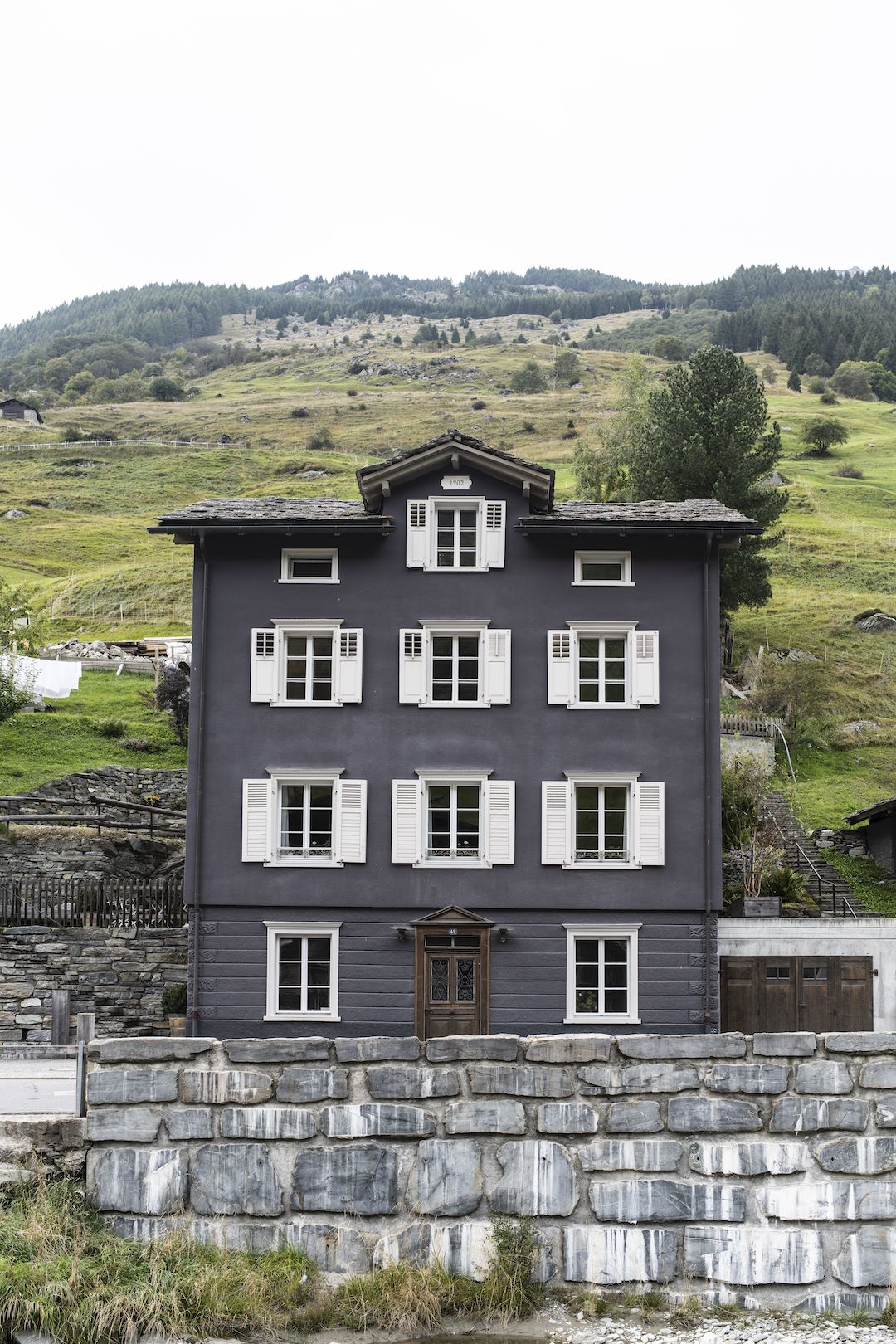 Brücke 49 guesthouse in Vals, a charcoal 1902 house with white shutters and door number 49 against a green hillside.