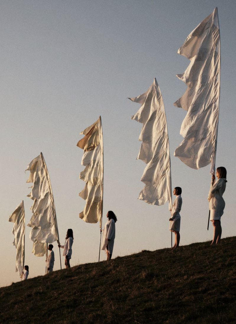 Commune Botanicals campaign image of figures in white carrying tall ivory flags across a Somerset hillside at dusk, sustainable natural self-care by Kate Neal and Rémi Paringaux, Bruton England | The Aficionados