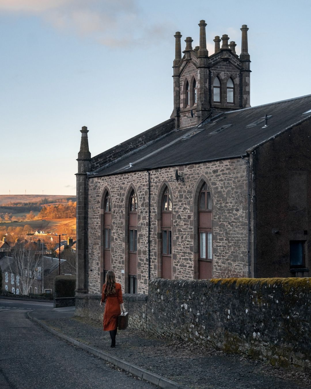 A woman in a rust-orange dress walks past The Church Tower, (Guesthouse) a converted Gothic stone church with tall arched windows, in the whisky town of Crieff, Perthshire.
