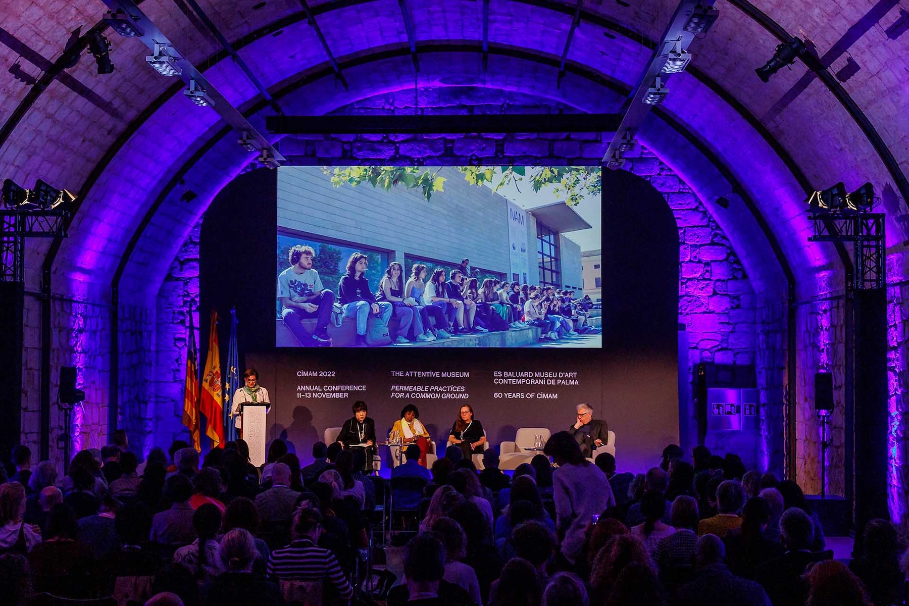 A full audience beneath the barrel-vaulted stone ceiling of the Aljub, lit blue for a conference panel and screen | The Aficionados