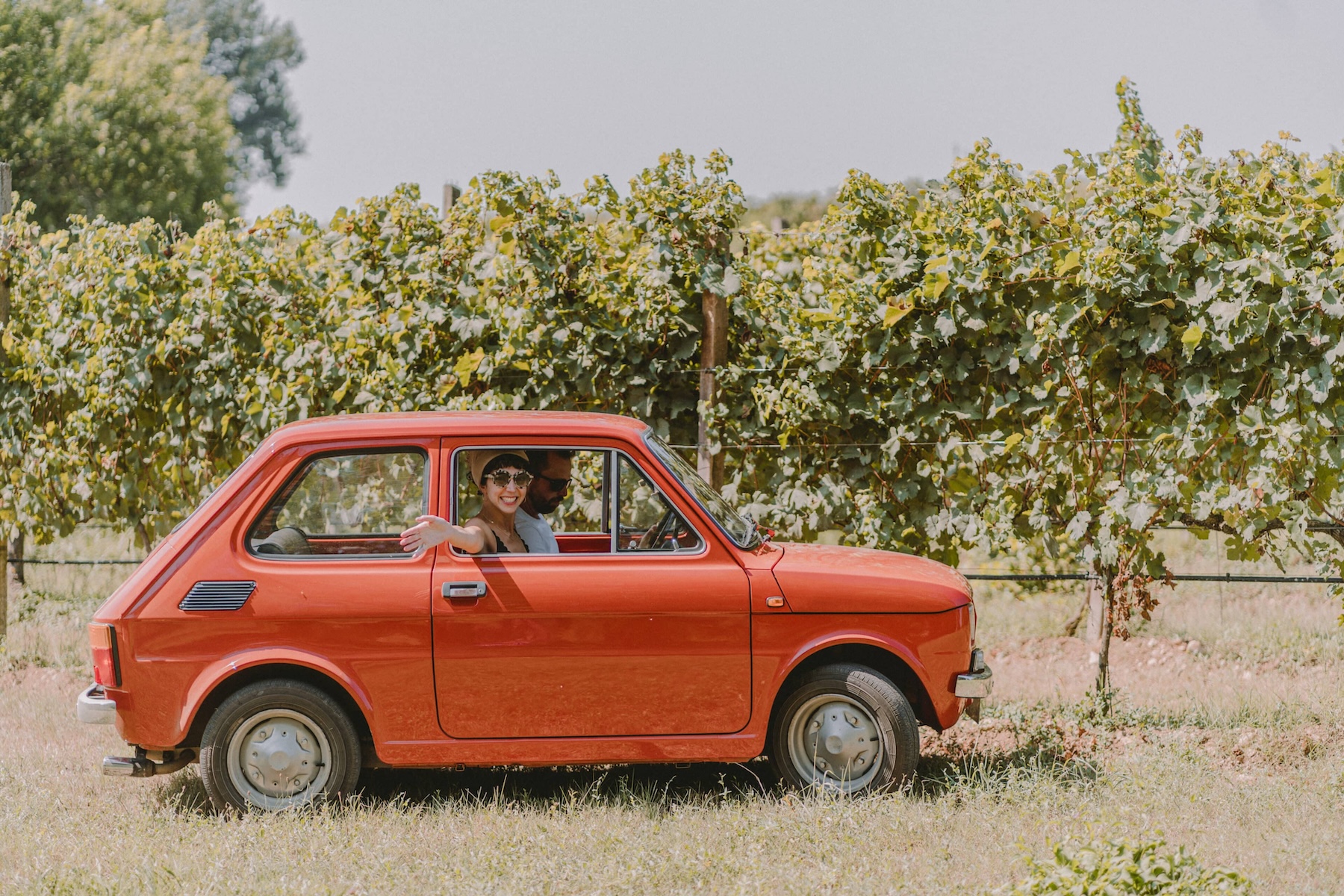 Smiling couple in a vintage red Fiat 126 parked among the vines of a northern Italian vineyard