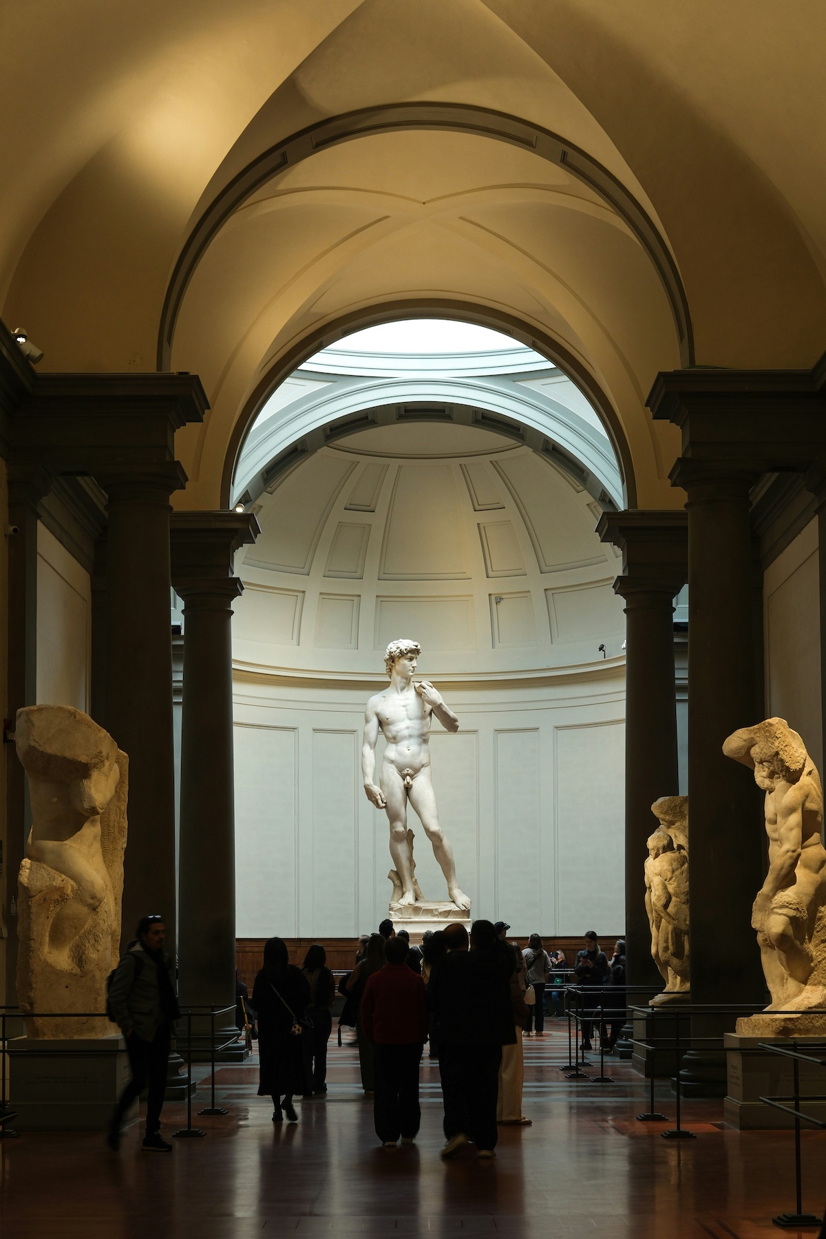 Michelangelo’s David beneath the skylit dome of the Tribune at the Galleria dell’Accademia, Florence, with his unfinished Prisoners lining the gallery and visitors gathered below.