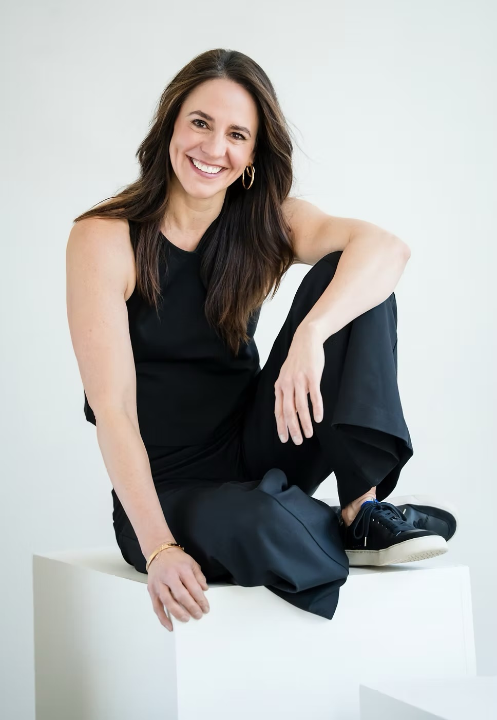 Dr Courtney Conley, founder of Gait Happens and author of 'Walk', with long brown hair and gold hoop earrings, seated in a black sleeveless top and smiling against a soft pink backdrop.