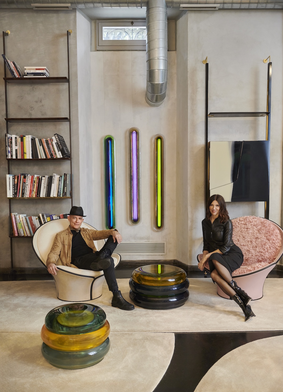 Draga & Aurel seated in sculptural armchairs below three glowing resin wall lights, with a stacked resin side table between them | The Aficionados