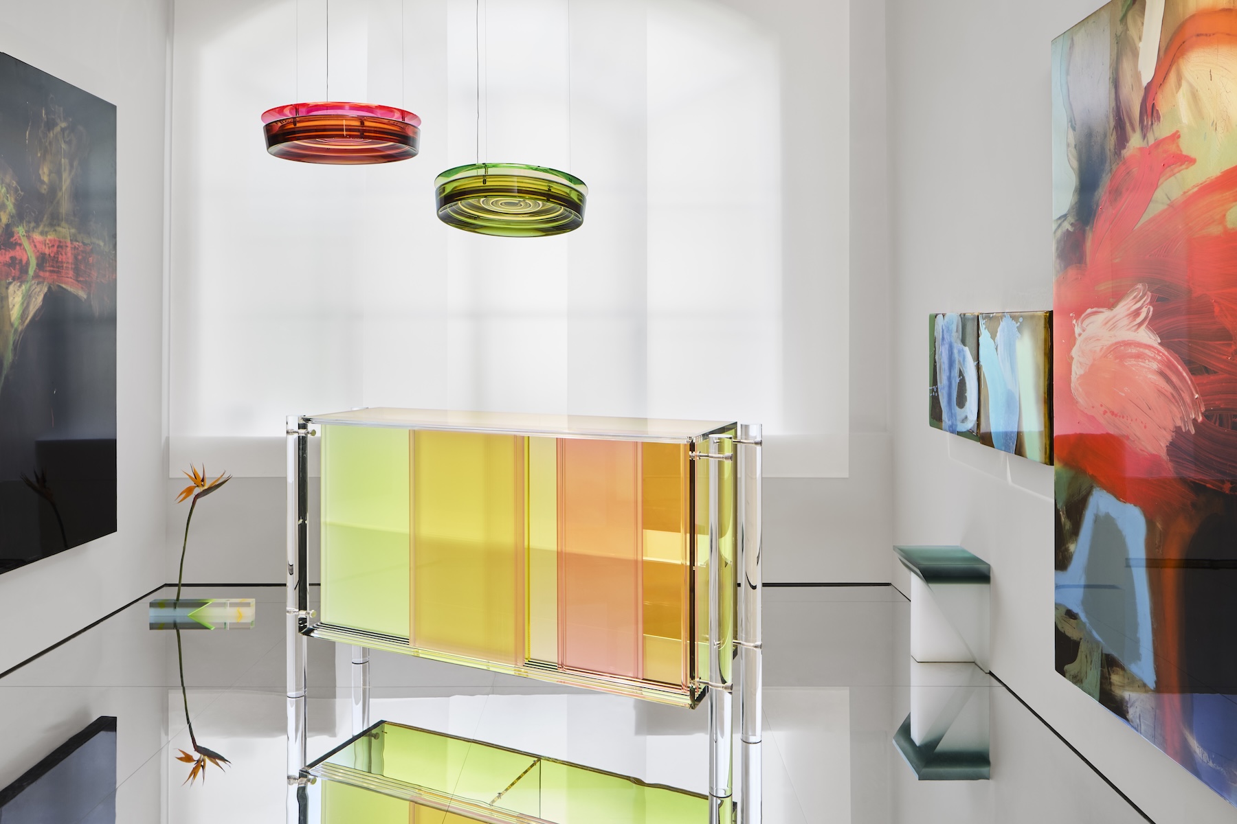 A transparent cabinet in green, yellow and pink beneath two coloured resin pendant lights, flanked by abstract paintings | The Aficionados