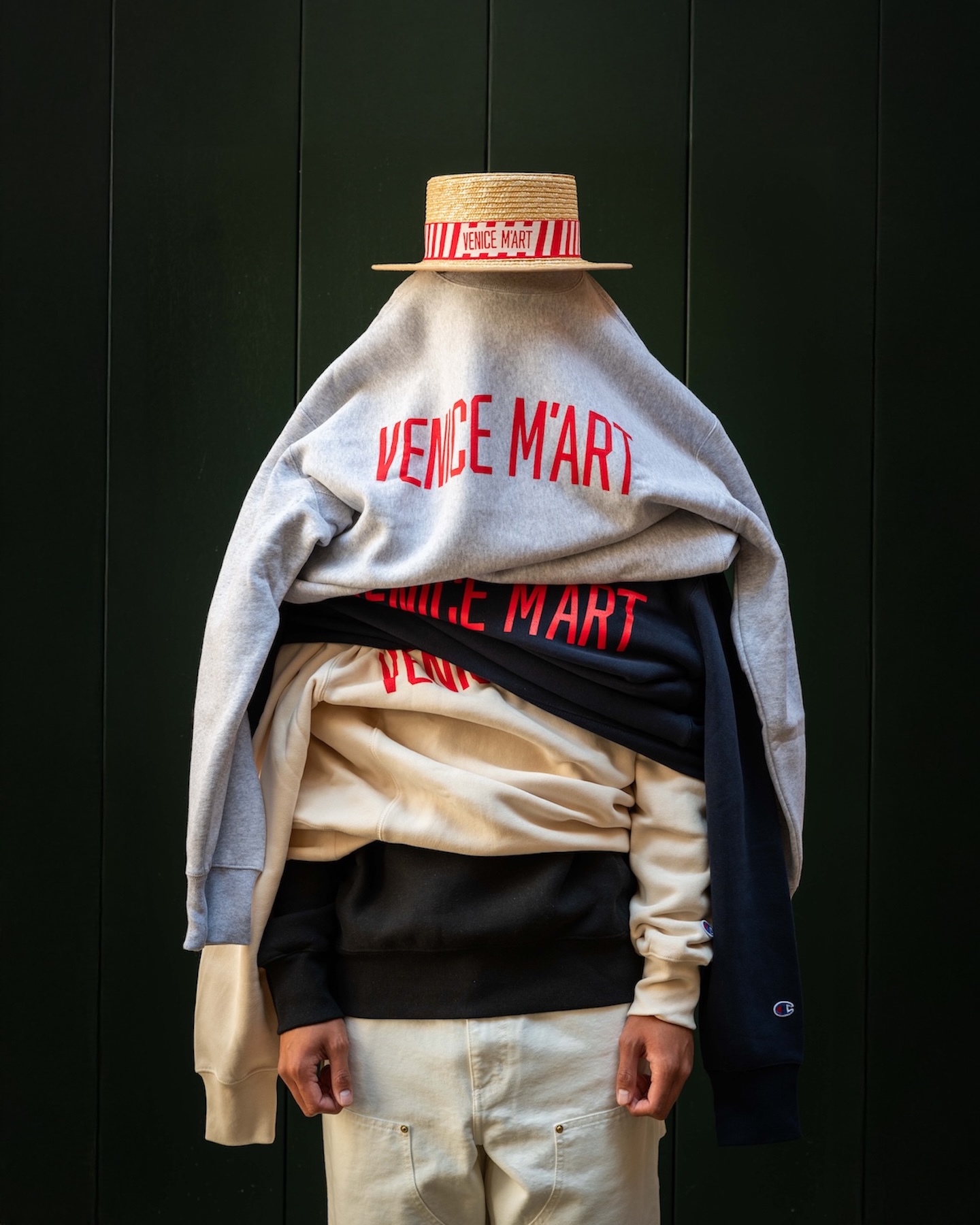 A person layered in a stack of Venice M'Art sweatshirts in grey, navy, cream and black, topped with a straw boater hat banded in Venice M'Art ribbon, against a dark green panelled wall.