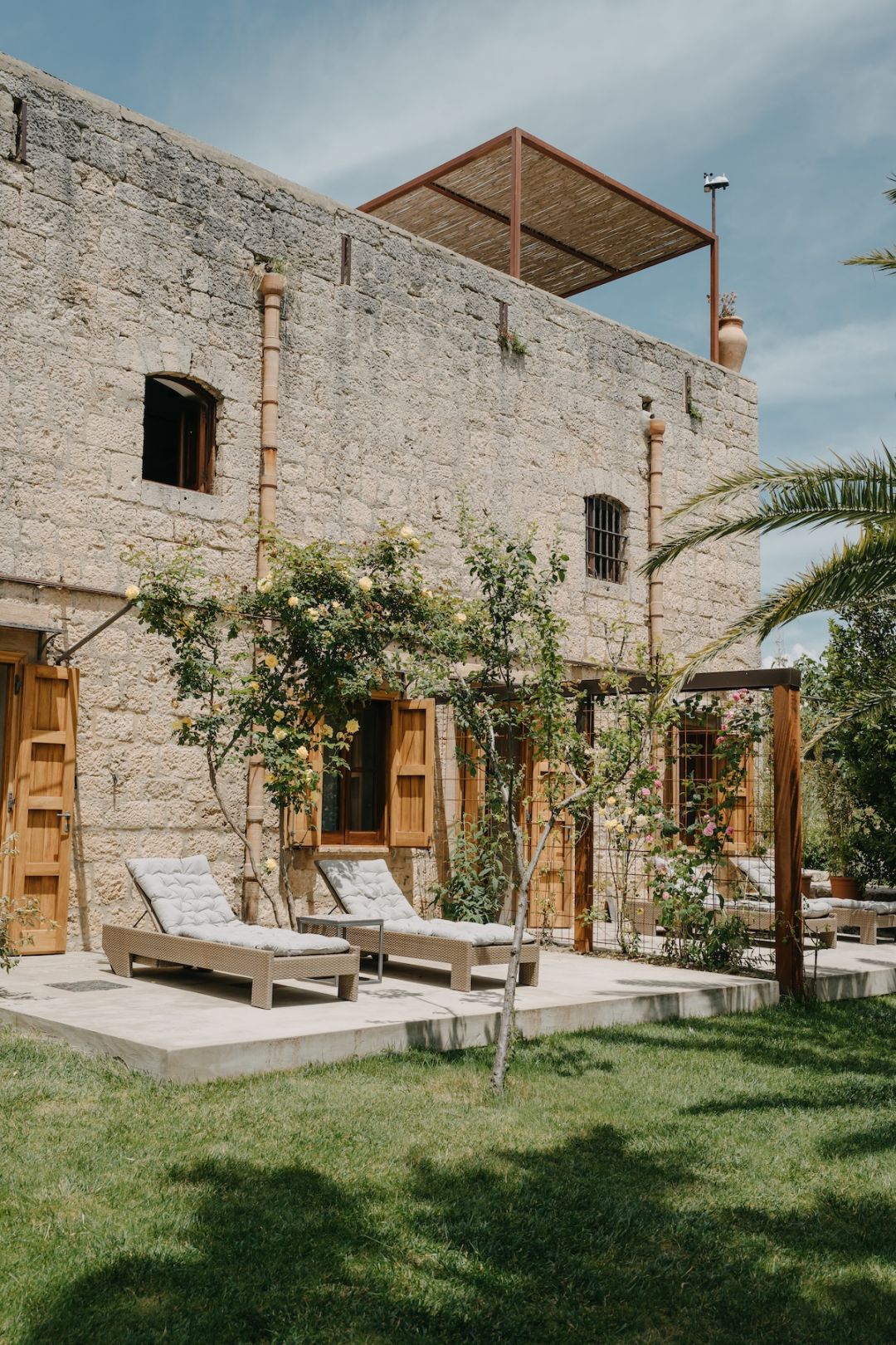 Pale-stone walls and a rooftop reed pergola at Susafa, a working Sicilian masseria, with climbing roses and sun loungers below.
