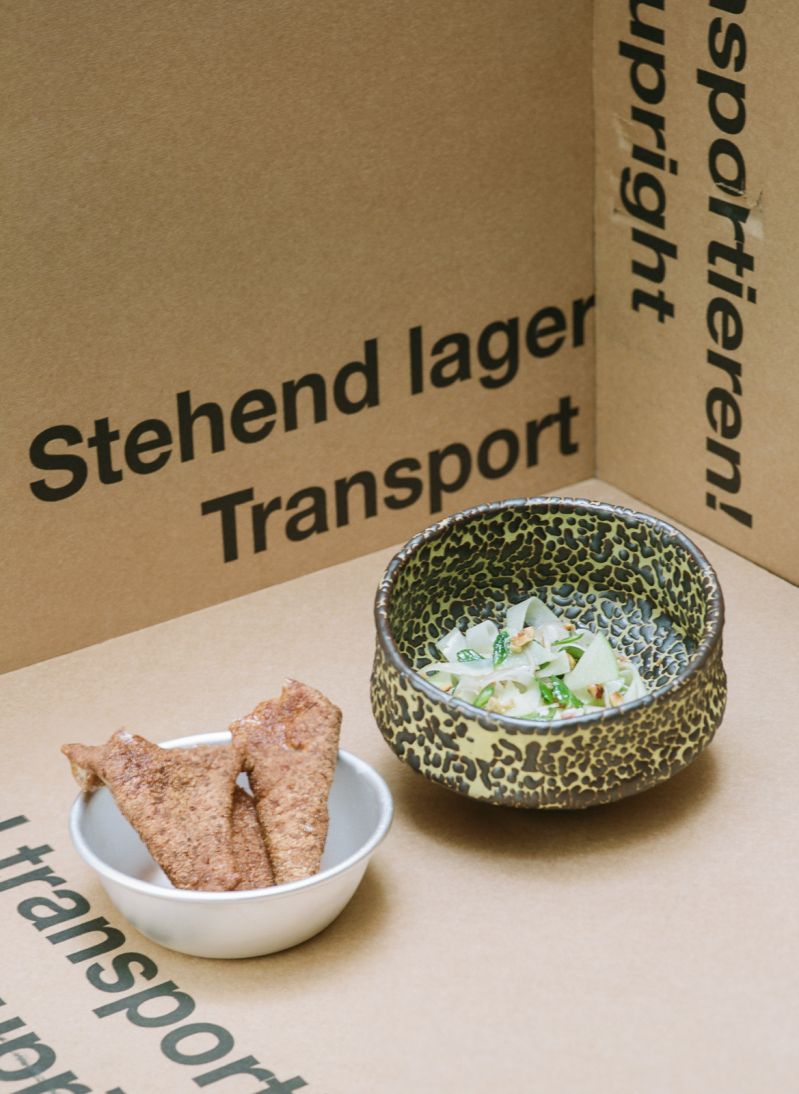 A speckled ceramic bowl of seasonal salad with crackers, plated on cardboard transport boxes in the playful style of Mraz & Sohn
