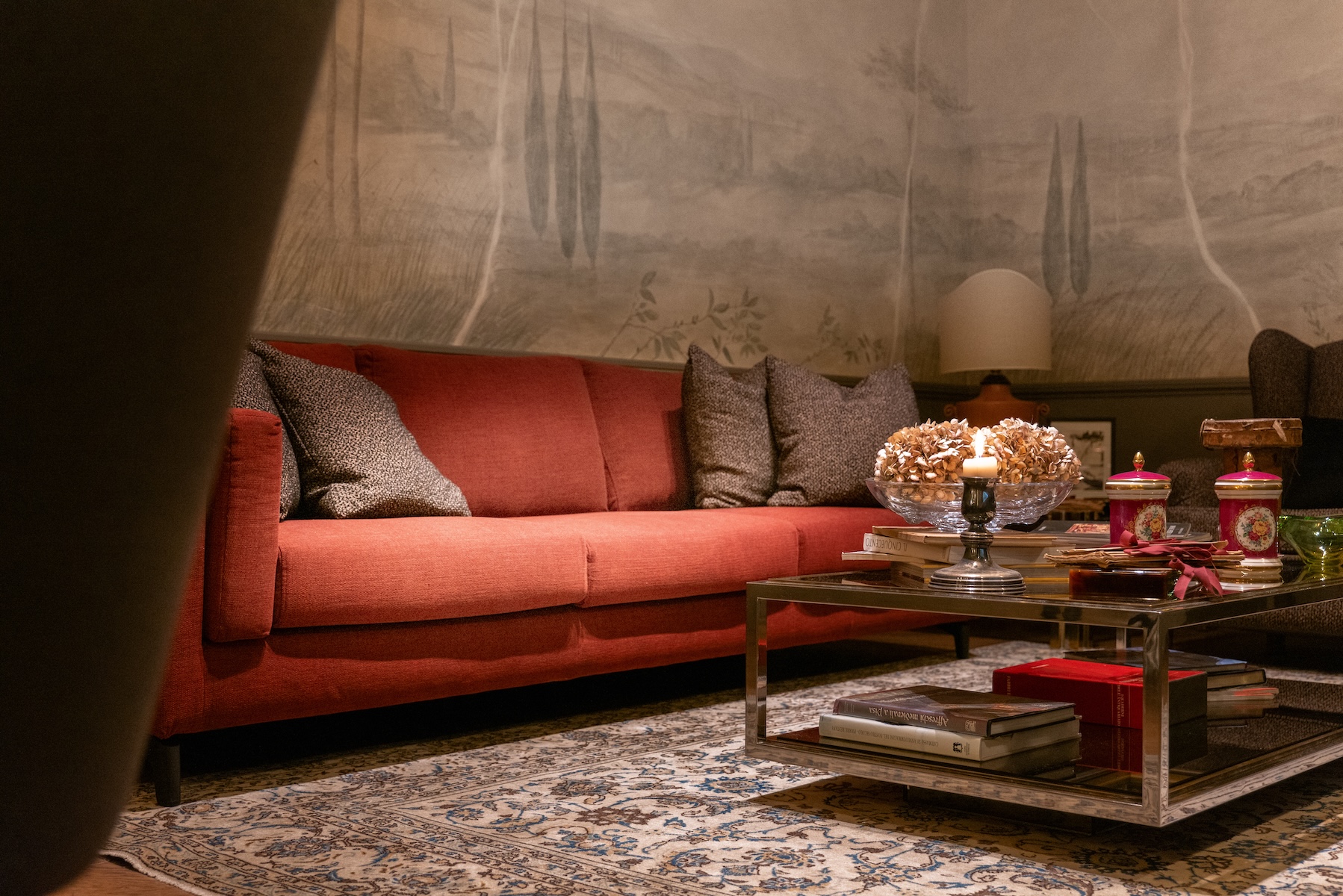 The living room at Lungarno Guicciardini 9, a brick-red sofa before a misty painted landscape of cypresses and Tuscan hills, with a glass and chrome table and an ivory and indigo rug. © Tommaso Pini