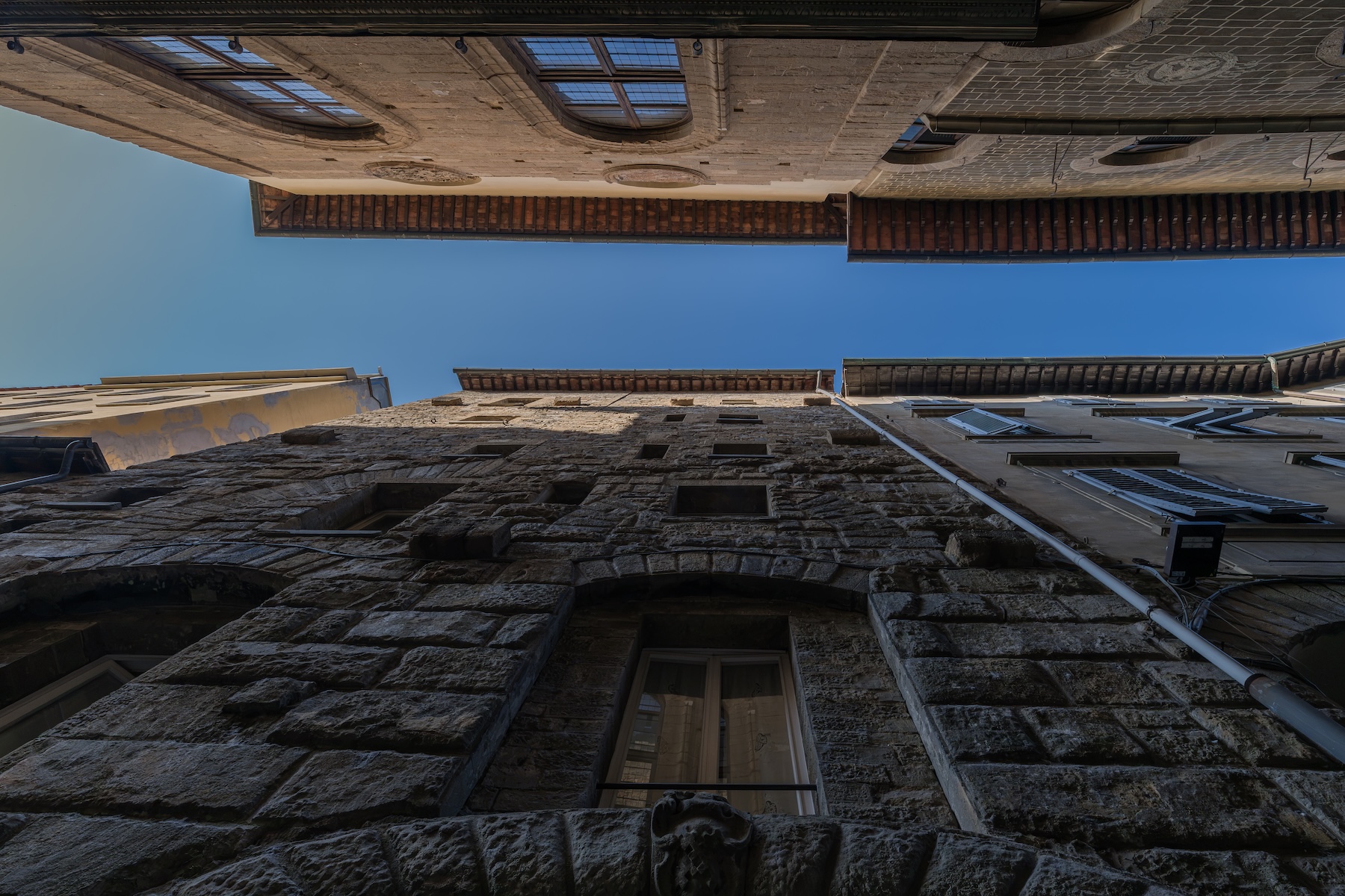 Looking up between medieval stone tower-houses in a narrow Florentine street, rough-hewn walls and small windows rising to overhanging timber eaves and a strip of blue sky.