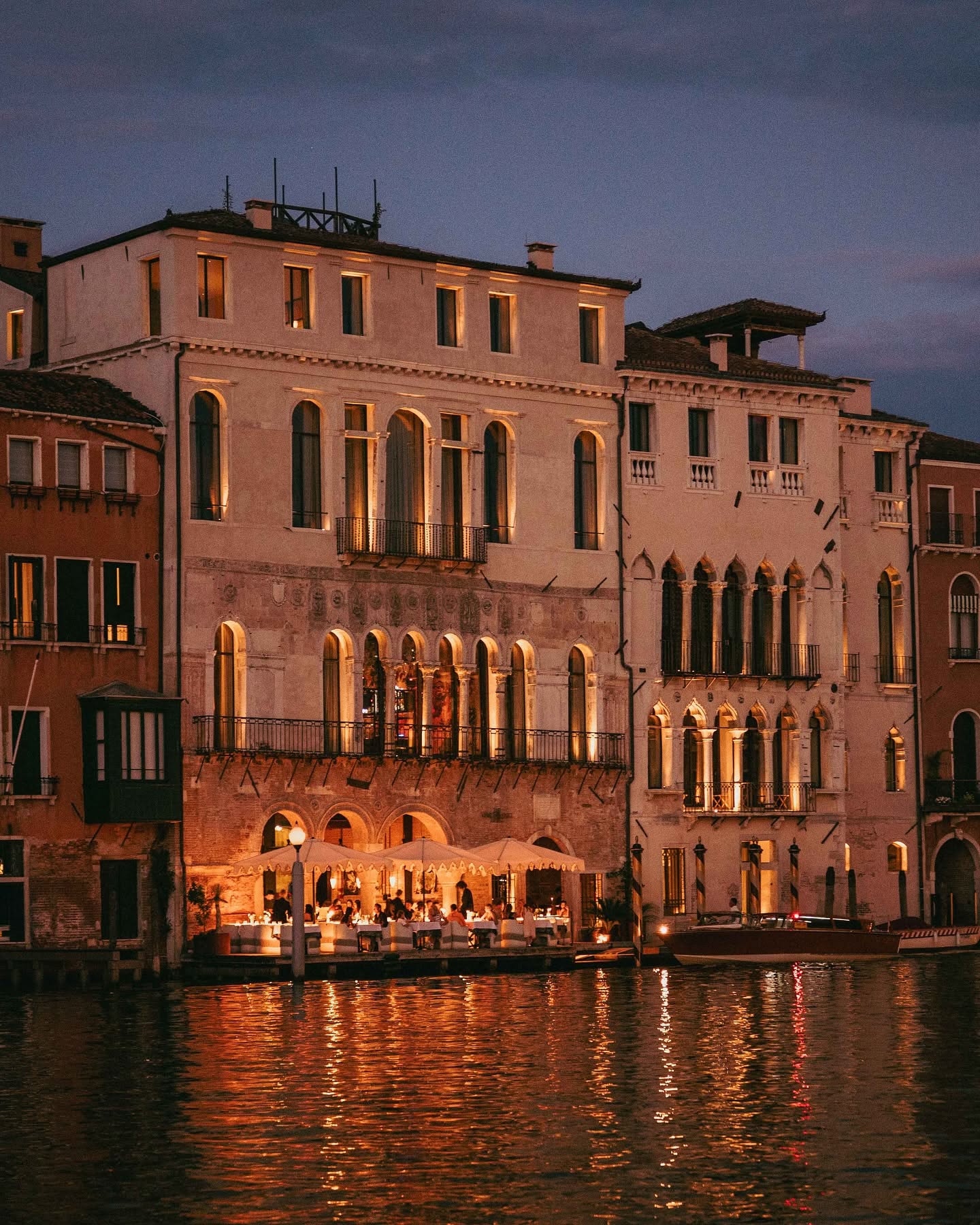 The Venice Venice Hotel lit at dusk on the Grand Canal, with Francy's terrace dining beneath white umbrellas at the water's edge.