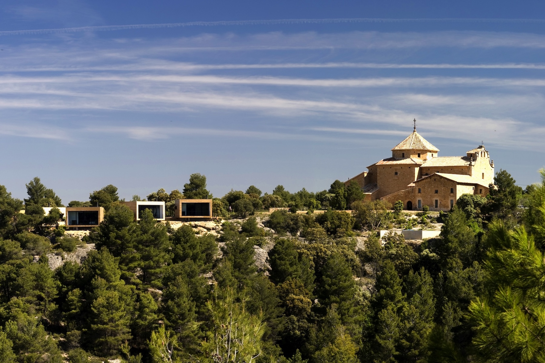 Timber and glass cubes stepped along the pine-covered ridge below the sixteenth-century hermitage | Consolación, Matarraña, Teruel, Spain | The Aficionados