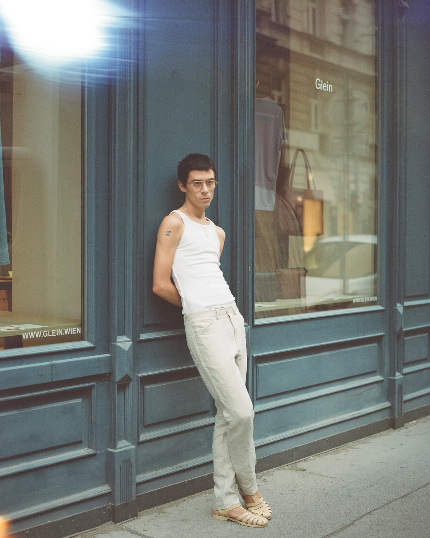 A man leaning against the blue-grey facade of the Glein store in Vienna, name and web address glein.wien lettered on the window