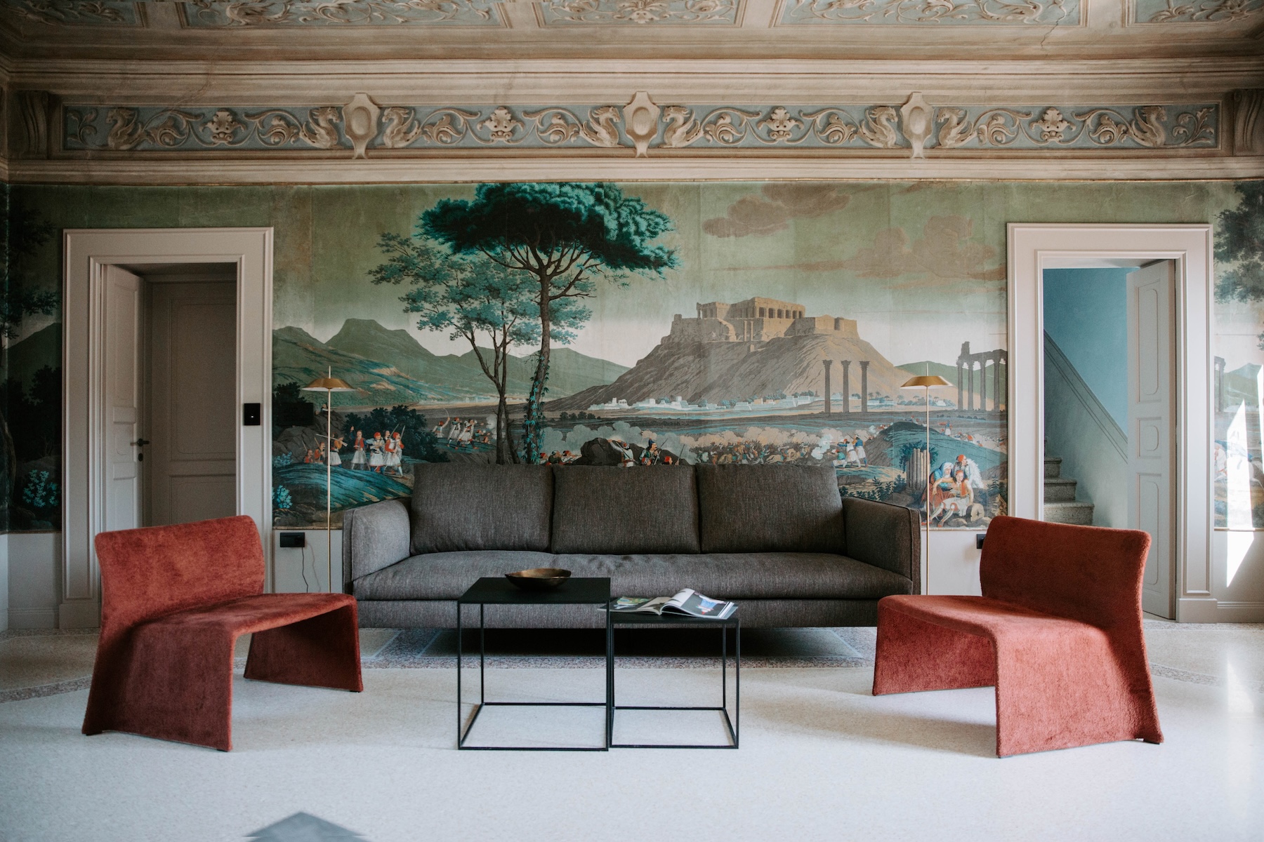 A salon at Relais Roncolo 1888 in Emilia-Romagna, Italy, its wall painted with a panoramic view of the Acropolis, set with a grey sofa and rust-coloured armchairs beneath a carved frieze.