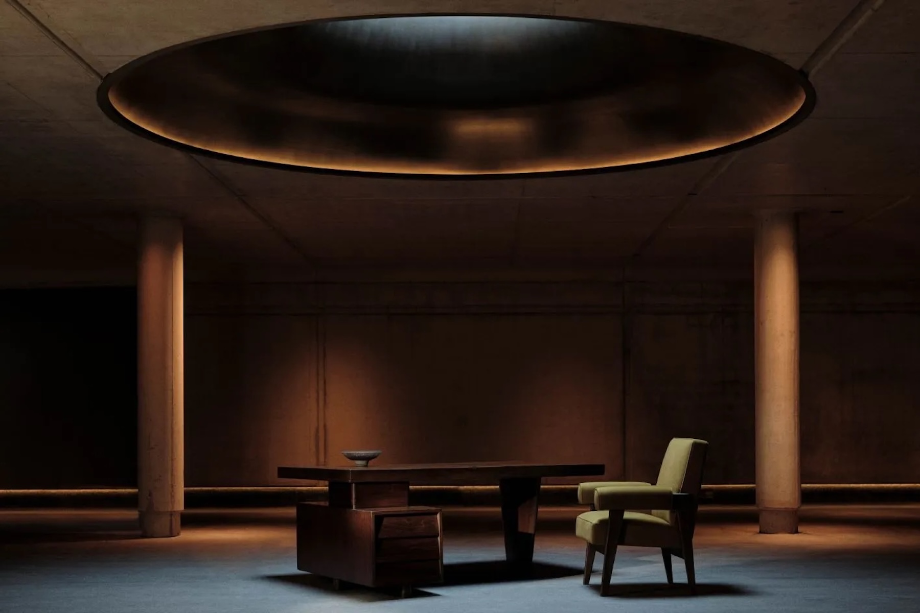 A timber desk and olive armchair beneath a glowing oval recess in the ceiling of a pillared concrete hall designed by Shinichiro Ogata | The Aficionados