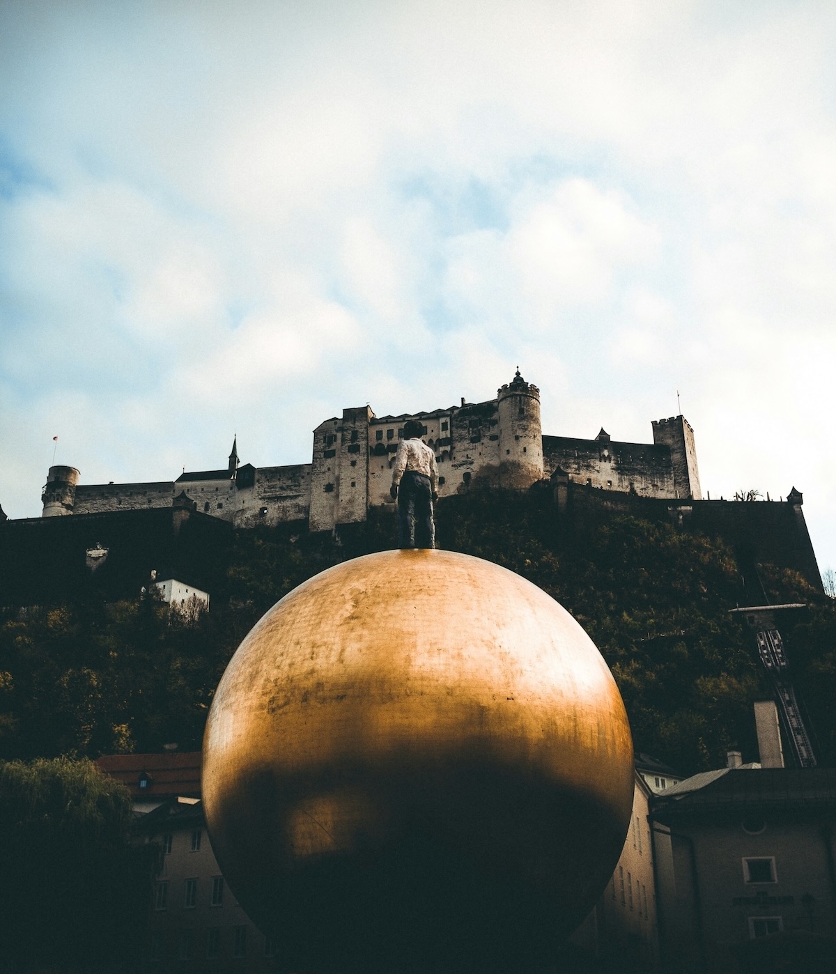 Stephan Balkenhol's Sphaera, a man standing on a golden sphere on Kapitelplatz beneath Hohensalzburg Fortress, Salzburg | The Aficionados