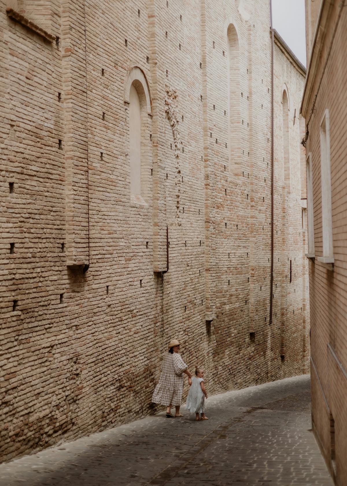 Mother and young child walking a quiet cobbled lane beside ancient brick walls in a medieval Le Marche hill town, Italy