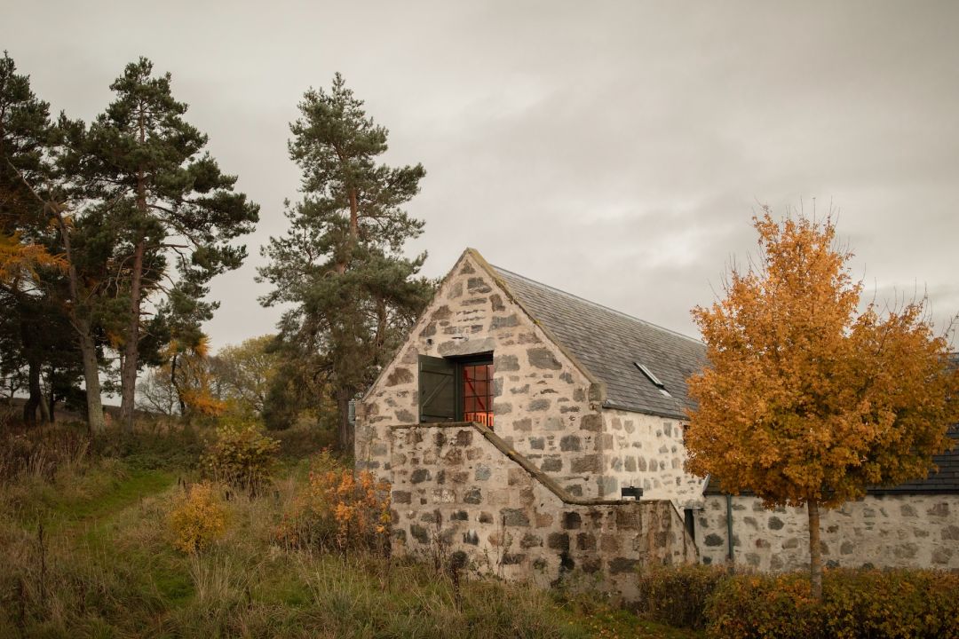 A converted stone steading at Killiehuntly Cottages in the Cairngorms, with a green shutter and red window, set among Scots pines and autumn trees.