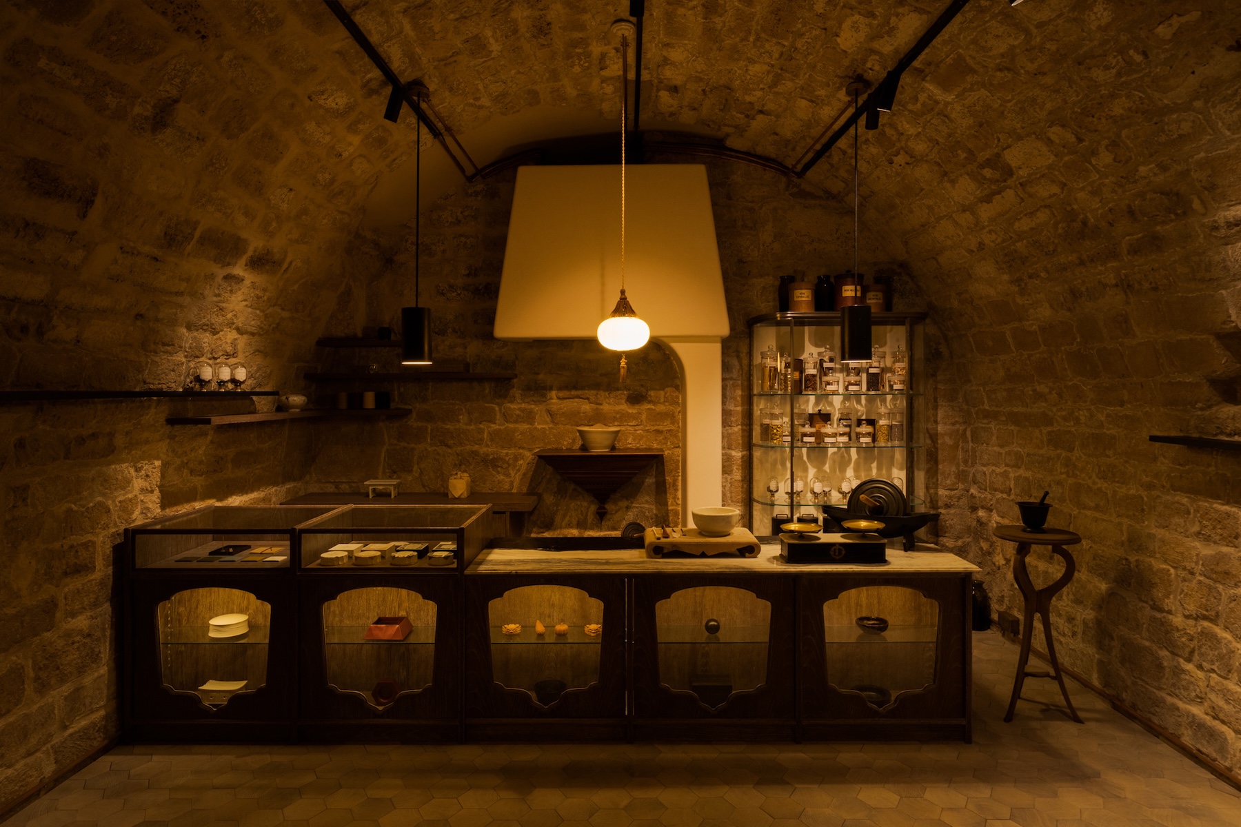 The vaulted stone cellar boutique at OGATA Paris, rue Debelleyme, with a dark timber counter of arched glass cases and a single pendant lamp | The Aficionados