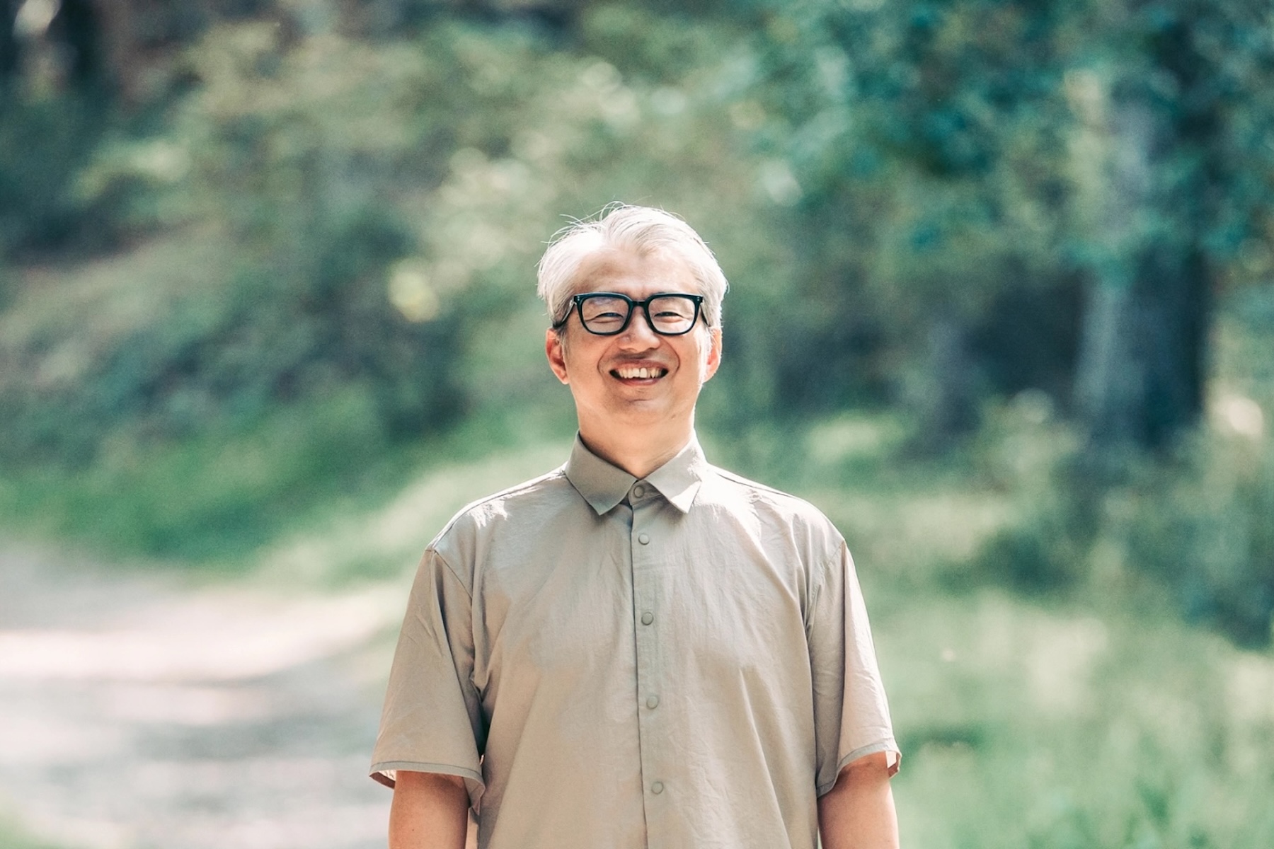 Takashi Fukuchi, founder and CEO of Stride, in black-framed glasses and a beige short-sleeved shirt, smiling on a sunlit woodland path surrounded by green foliage.