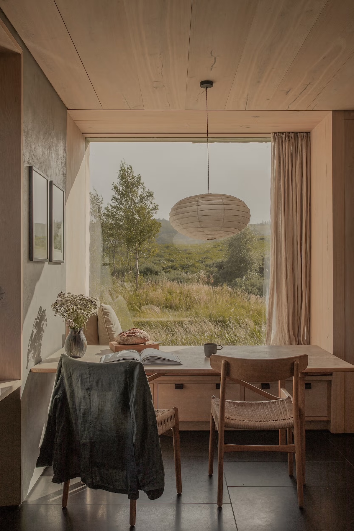 Scandi-Scot interior with an oak dining chair and linen throw beneath a paper lantern, beside a picture window onto Highland grassland, at a Wildland guesthouse in the Cairngorms.