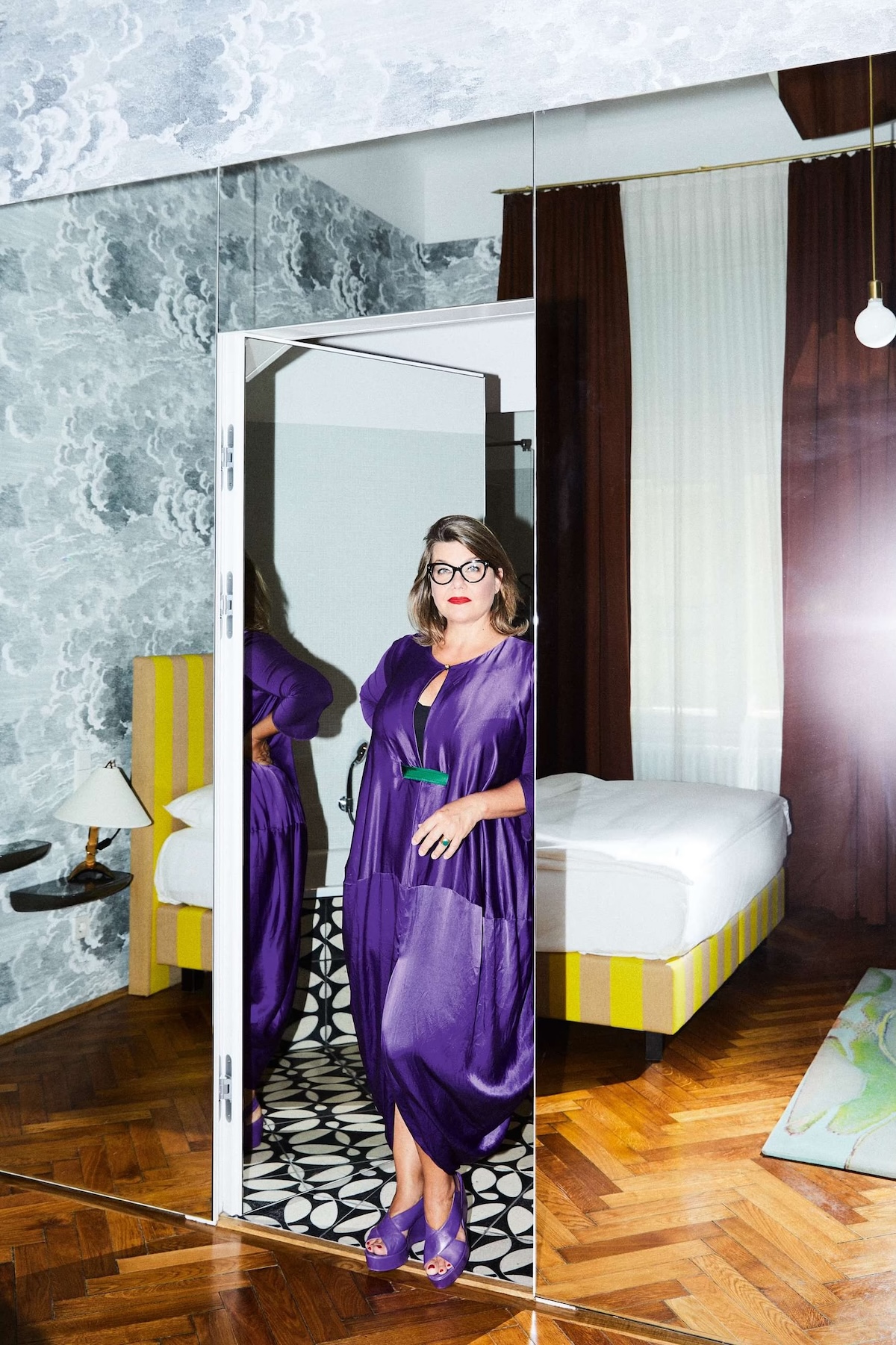 Lilli Hollein in a purple silk dress in the doorway of her cloud-wallpapered room at Hotel Altstadt Vienna, beside a yellow striped bed