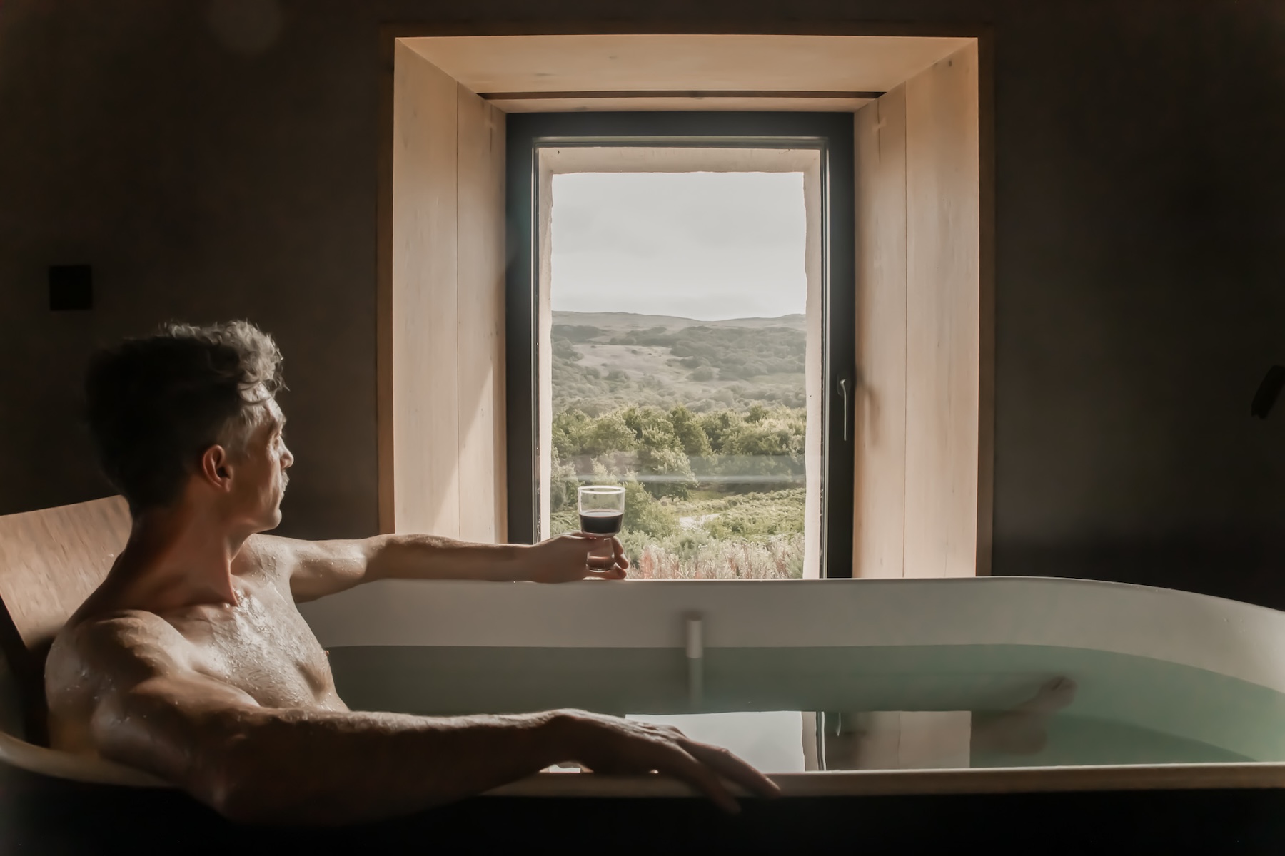 Man in a freestanding bath with a glass of red wine, looking through a deep oak-framed window to Highland hills and water at Kyle House, Kyle of Tongue, Sutherland. Filename: kyle-house-sutherland-bath-highland-view.jpg