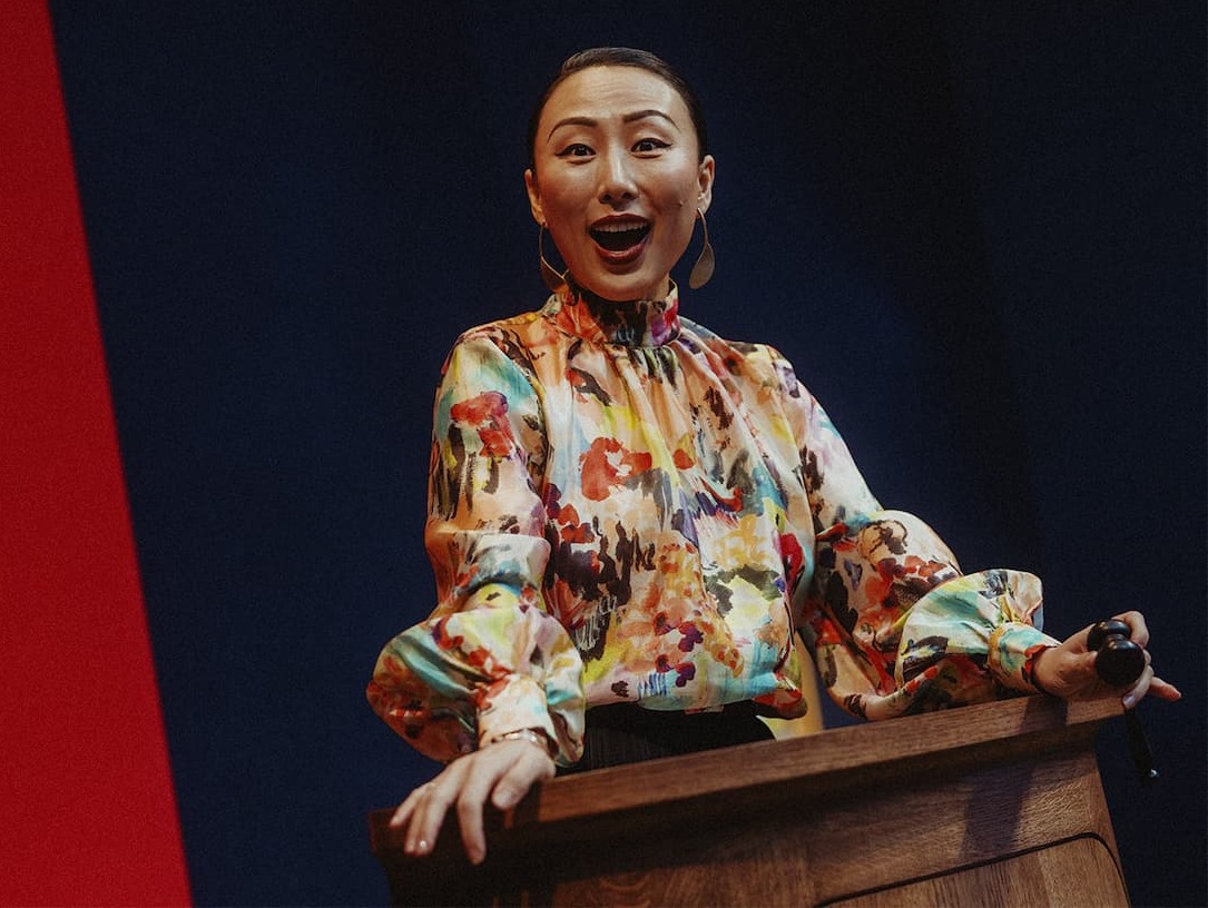 Christie's director and global auctioneer Yü-Ge Wang mid-speech at a wooden rostrum, holding a microphone in a floral-print blouse against a red and dark blue backdrop.