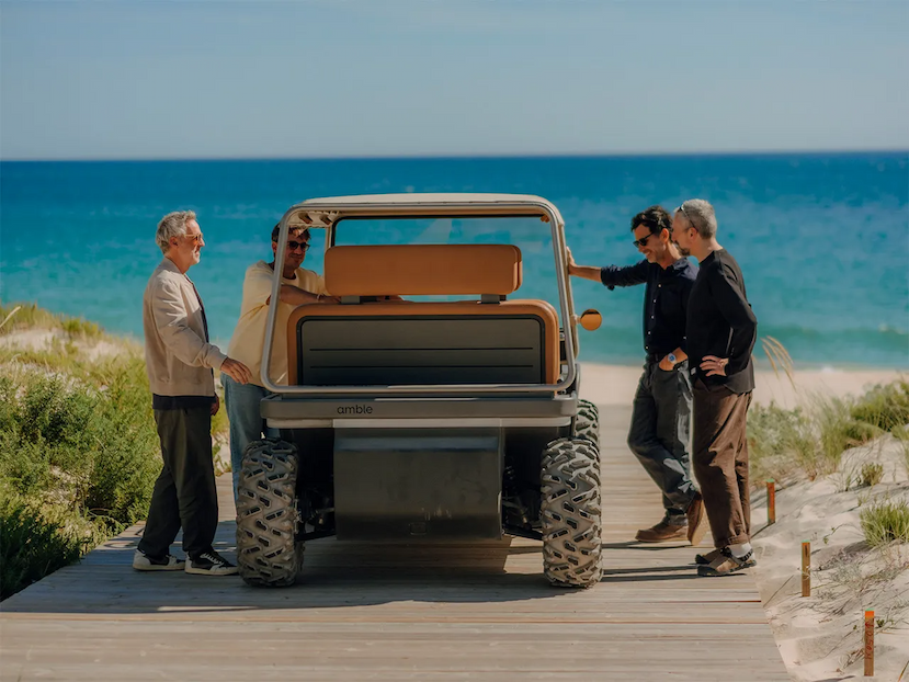 Amble co-founders beside the Amble One, a cork-wheeled electric four-seater buggy with chunky off-road tyres, standing on a timber boardwalk through dune grass with the Atlantic behind them.