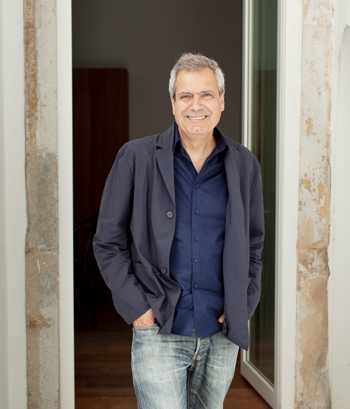 Portuguese architect Manuel Aires Mateus, founder of Aires Mateus, smiling in a navy shirt and jacket with his hands in his pockets, standing in a doorway framed by weathered plaster walls.