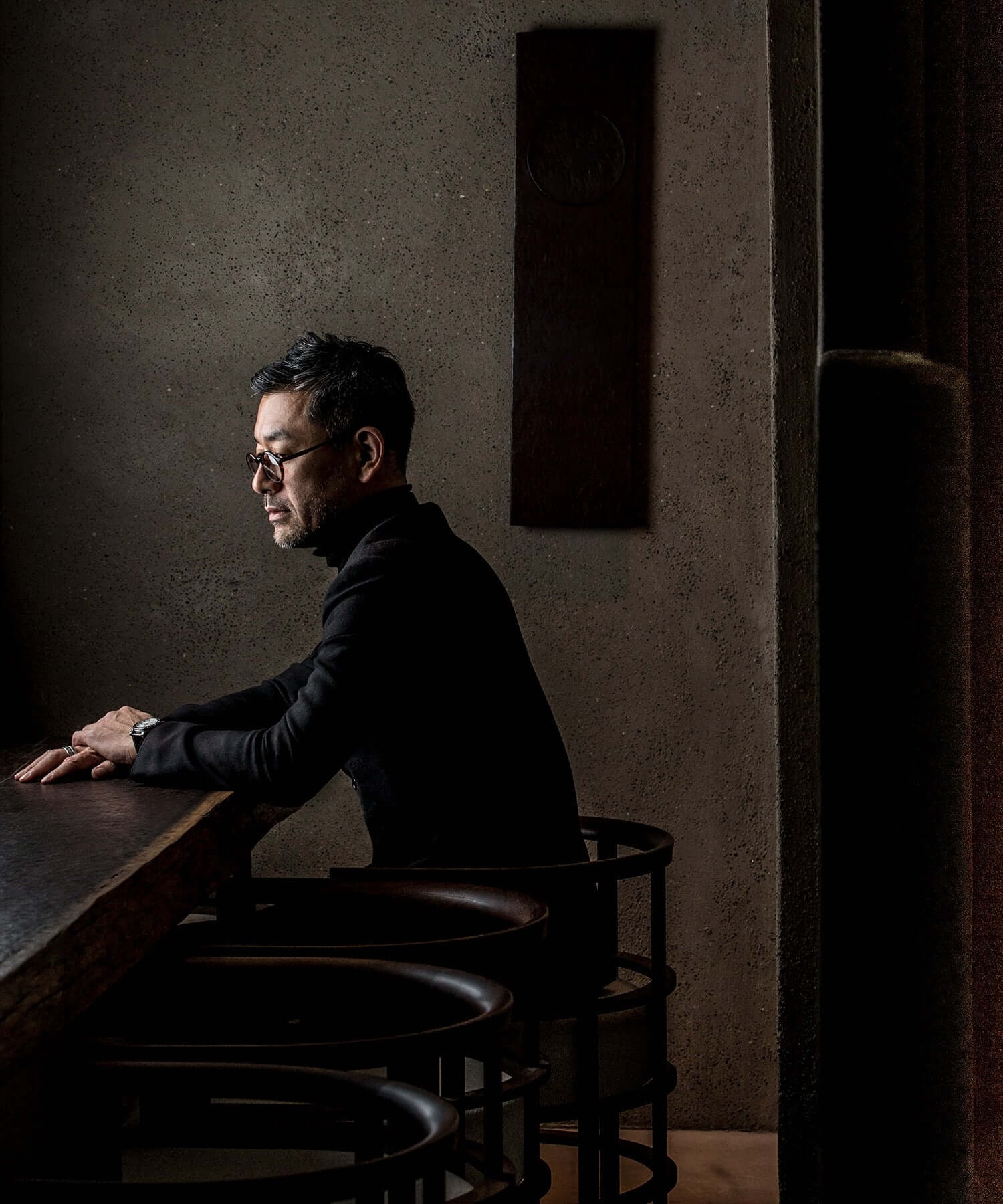 Shinichiro Ogata in profile at a dark timber counter, in a black roll-neck against a textured plaster wall | The Aficionados