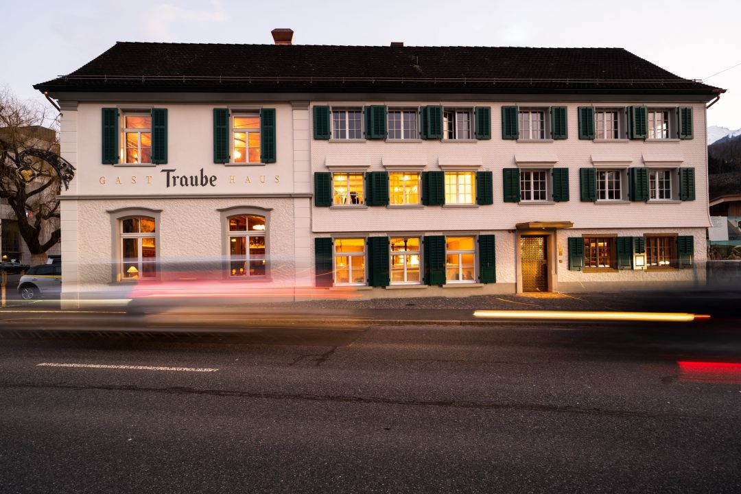 The Traube guesthouse in Buchs lit at dusk, white facade with green shutters and passing car light trails, Ostschweiz.