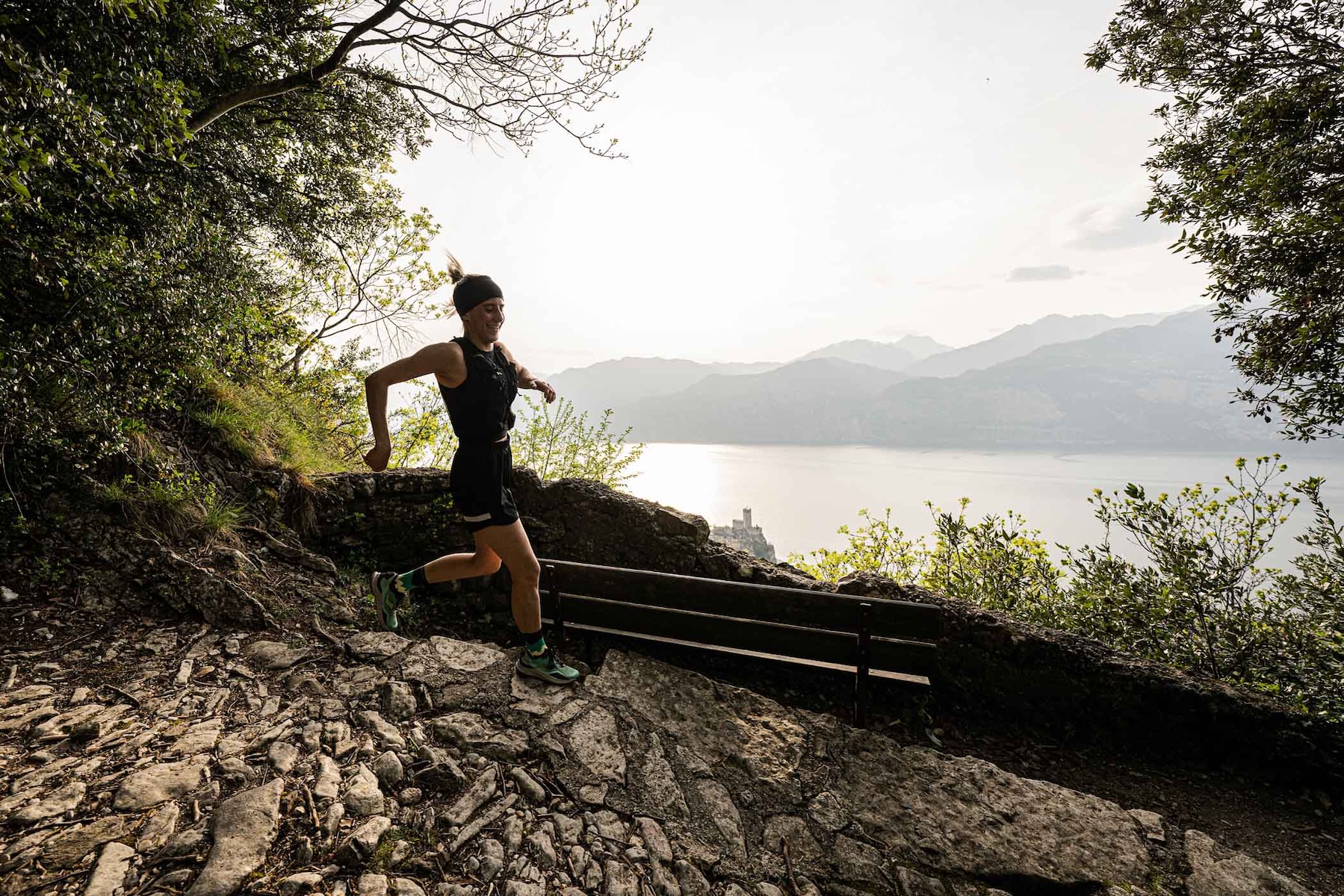 Runner on a rocky trail high above Lake Garda at sunset, the castle of Malcesine and the mountains catching the light below.