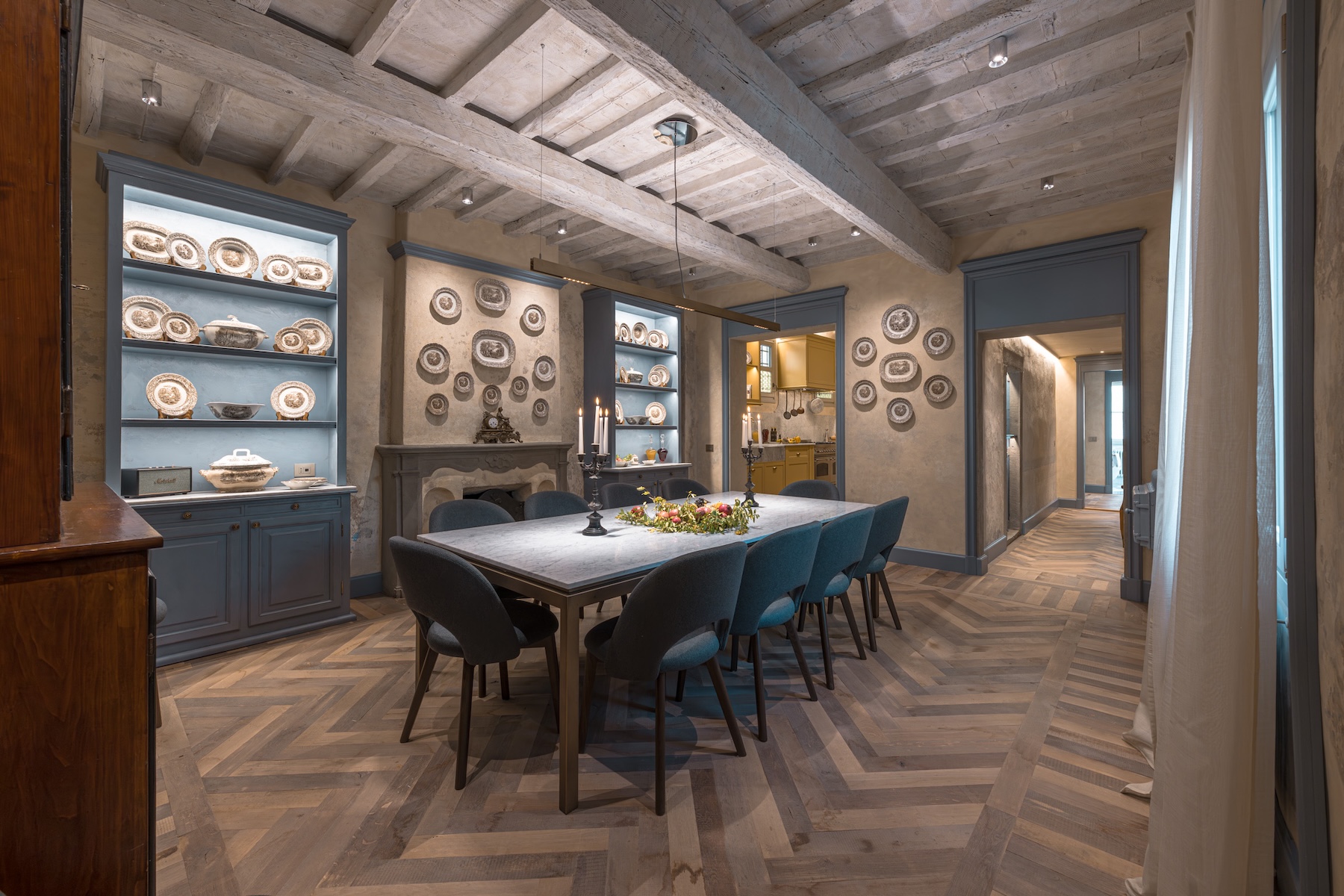 The dining room at Santa Croce Historical Home, a marble table for twelve beneath timber beams, blue-grey dressers of brown transferware, a stone fireplace and chevron oak floor leading to the ochre kitchen. © Tommaso Pini