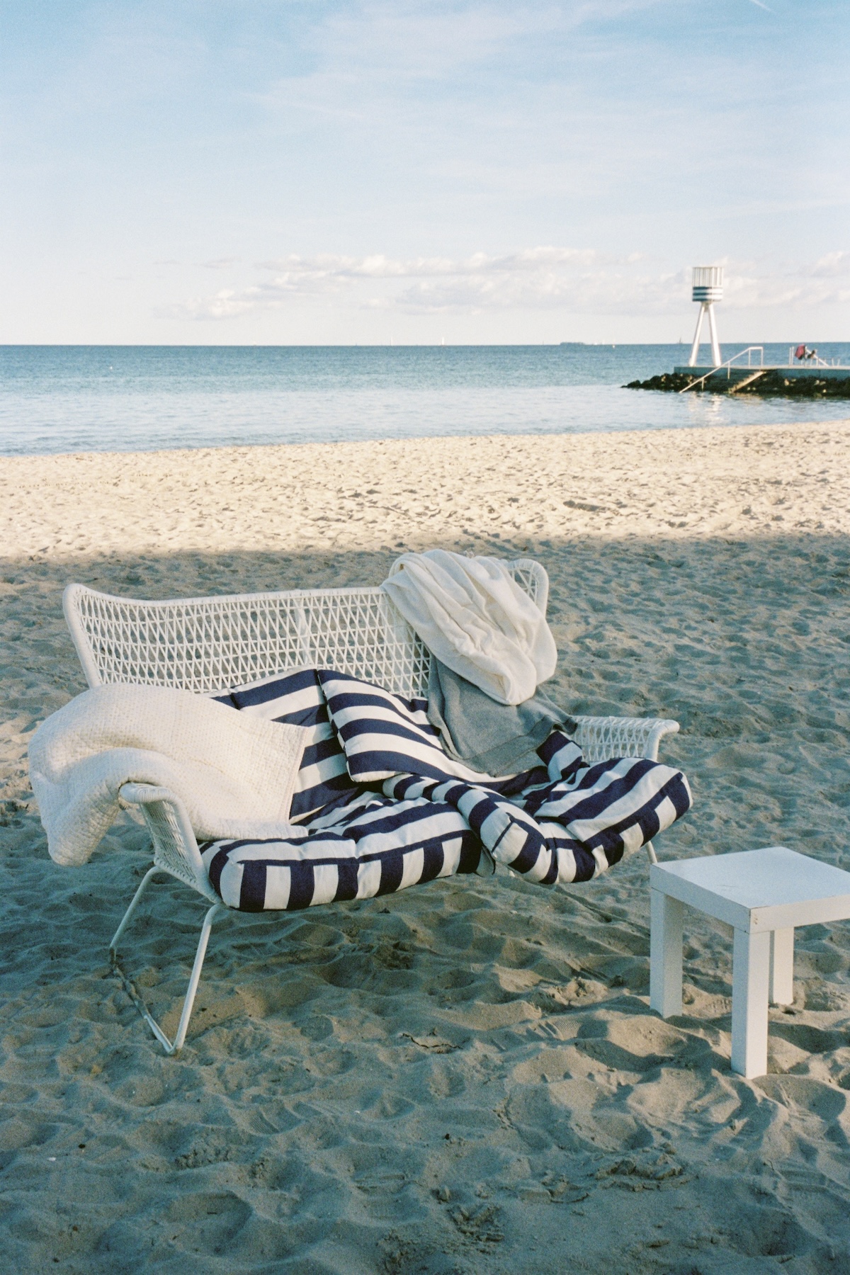 A white wicker chair with navy-striped cushions on Bellevue Beach, an Arne Jacobsen lifeguard tower on the shoreline beyond.