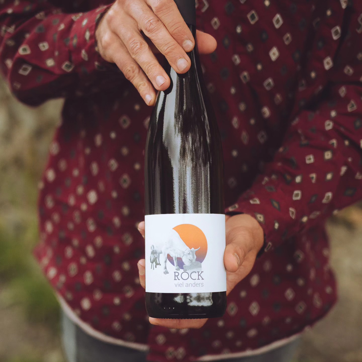 A bottle of Röck Viel Anders held against a patterned burgundy shirt, South Tyrol natural wine | The Aficionados