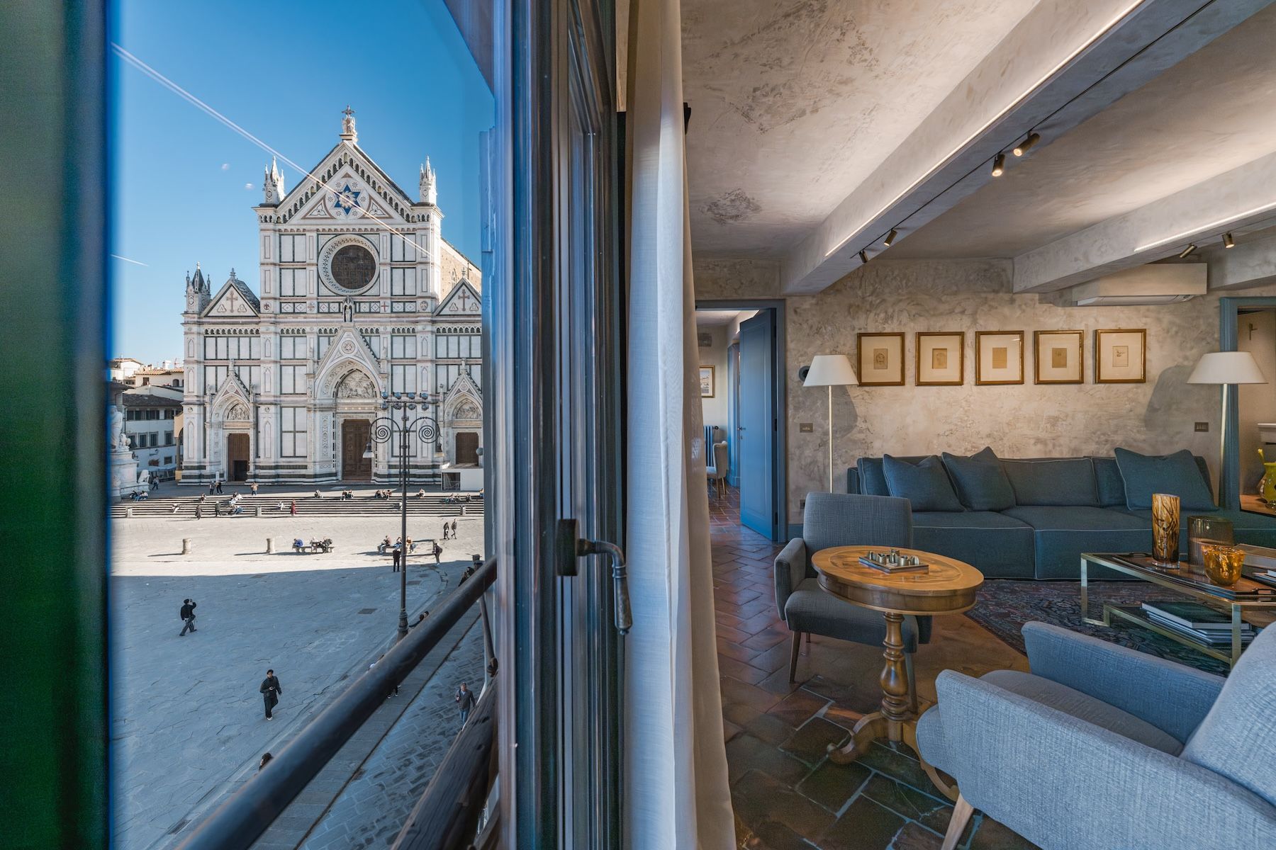 A window at Santa Croce Historical Home opening onto the Basilica of Santa Croce across the piazza, beside a living room of blue sofa, walnut games table and five framed drawings. © Tommaso Pini
