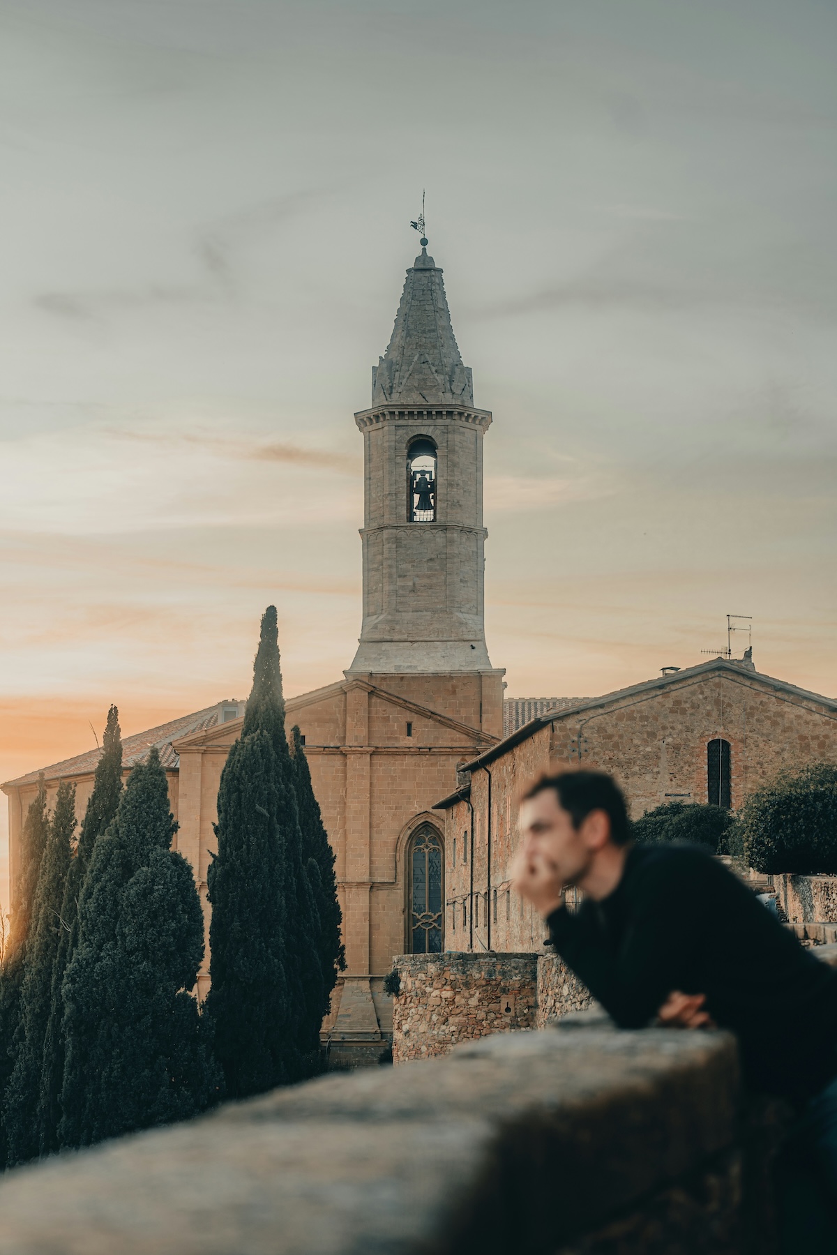 The bell tower of Pienza's Duomo rising above cypresses at dusk, with a visitor resting on the wall | The Aficionados