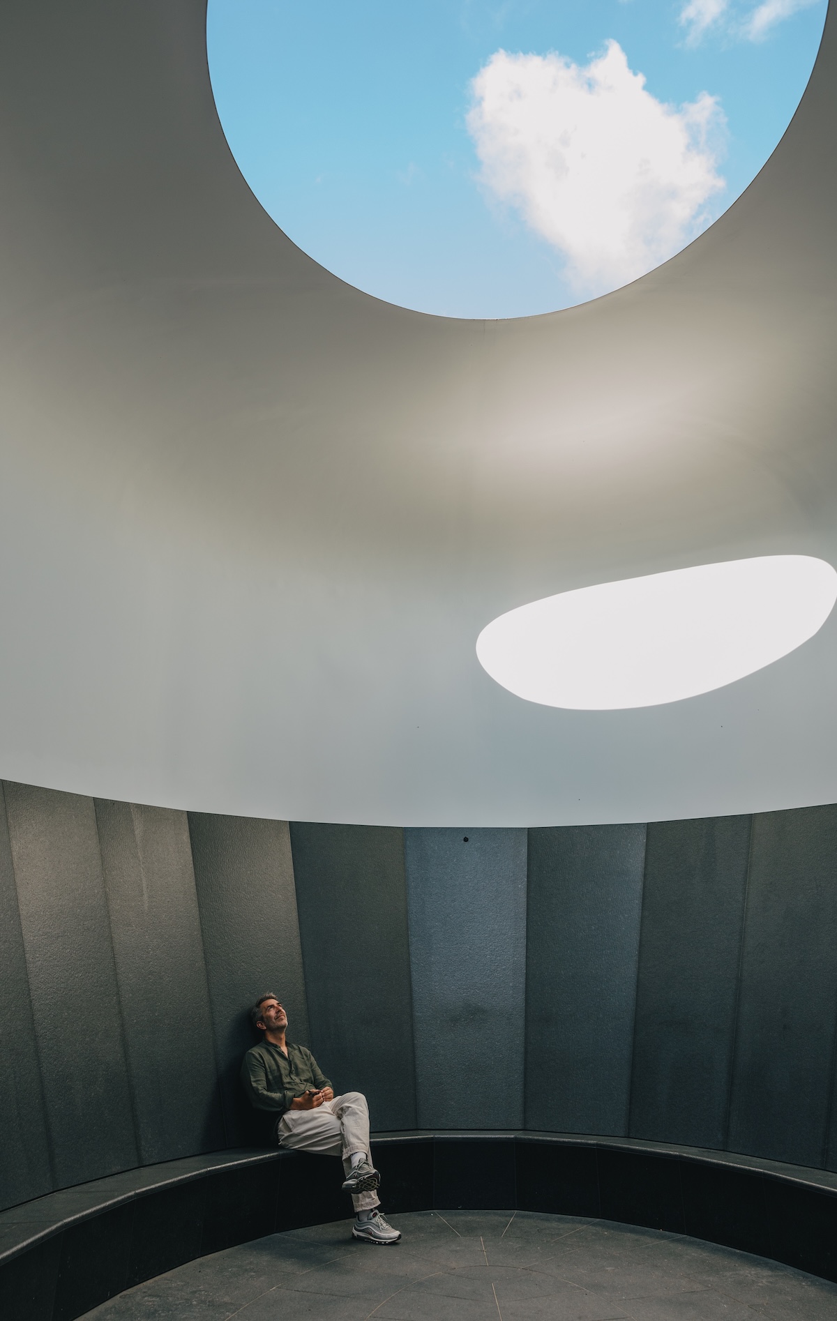 A visitor sits on the granite bench inside James Turrell’s Skyspace at Tannegg above Oberlech, looking up through the elliptical opening to a cloud in a blue sky | The Aficionados