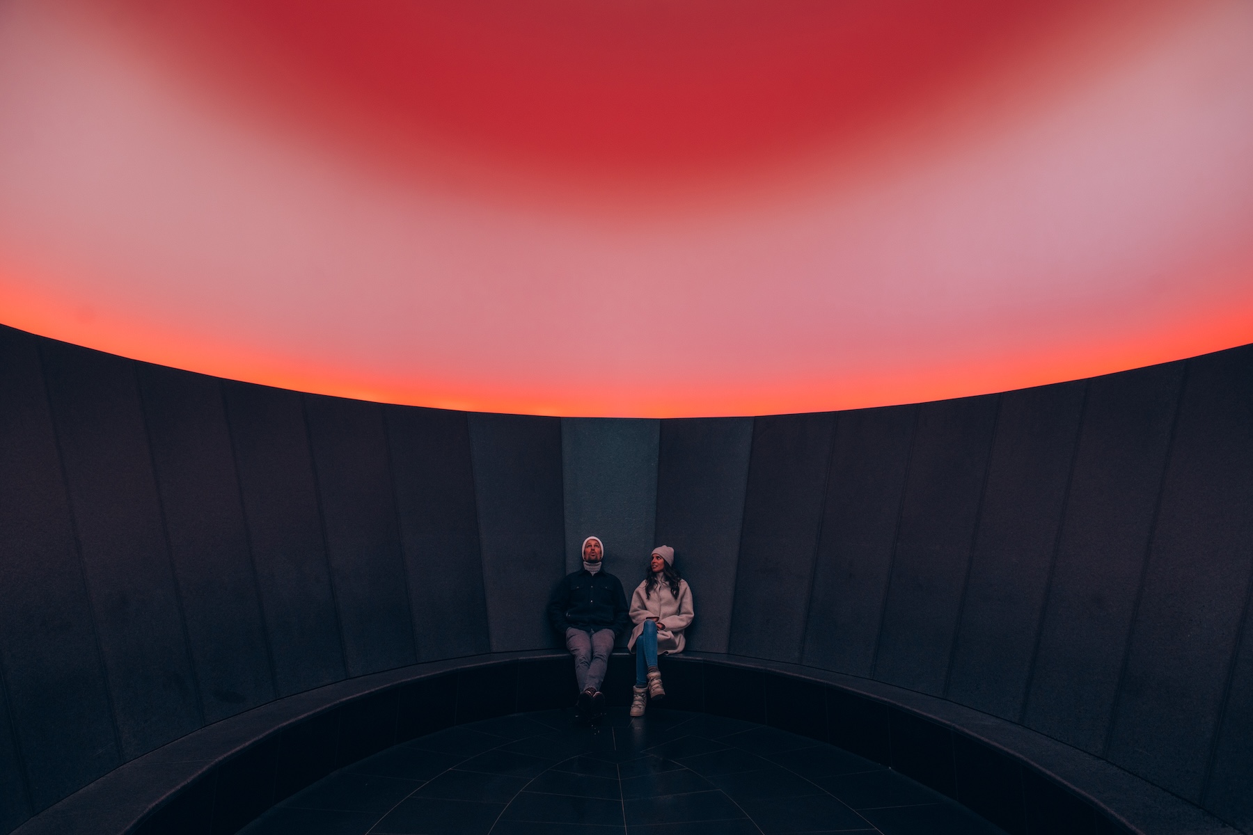Two visitors seated on the black granite bench of James Turrell’s Skyspace-Lech, looking up at an elliptical opening filled with red and pink light, Oberlech, Austria | The Aficionados