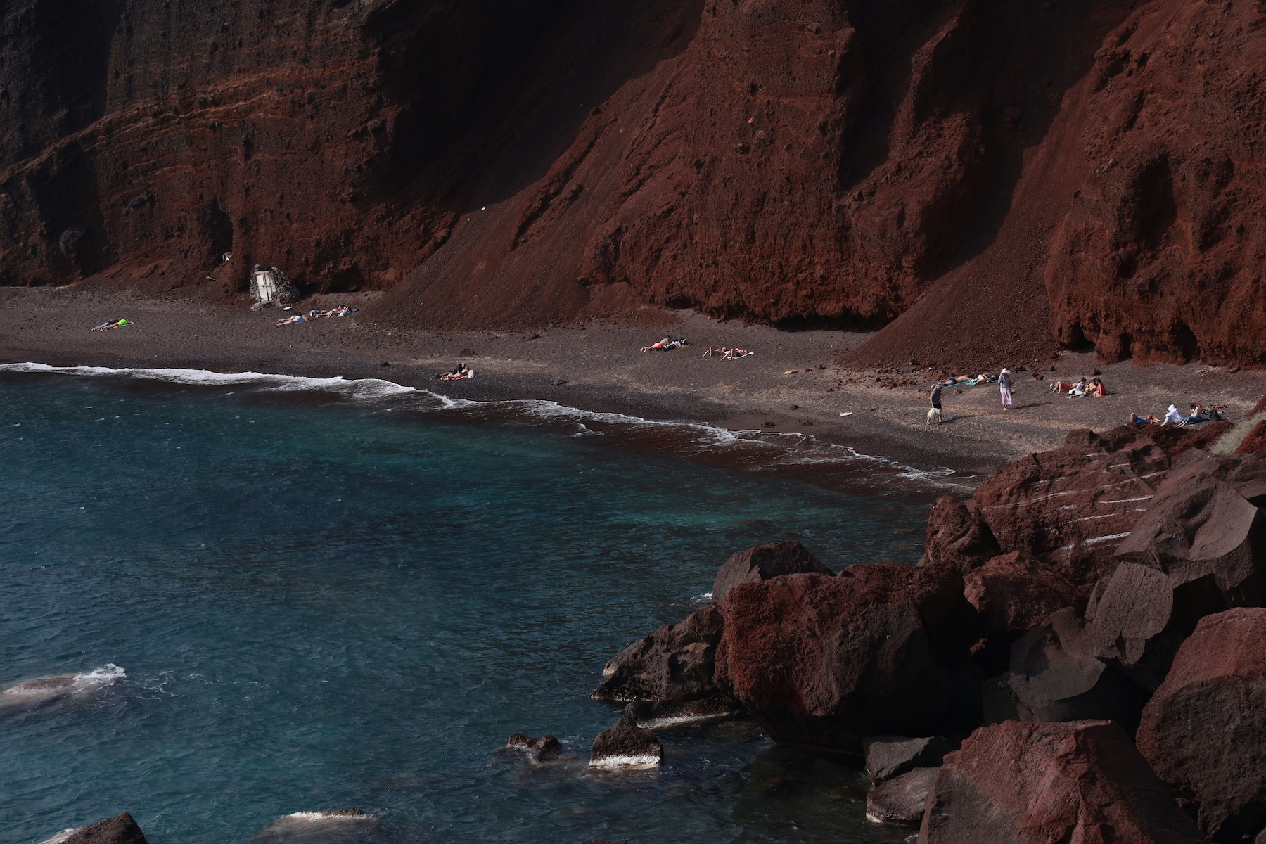 Spectacular Red Beaches > bay with poeple lying on the red sands backed by a steep red cliff |Santorini Noir: Volcanic Beauty, Architecture and Cycladic Soul| The Cyclades Island of Greece | The Aficionados 