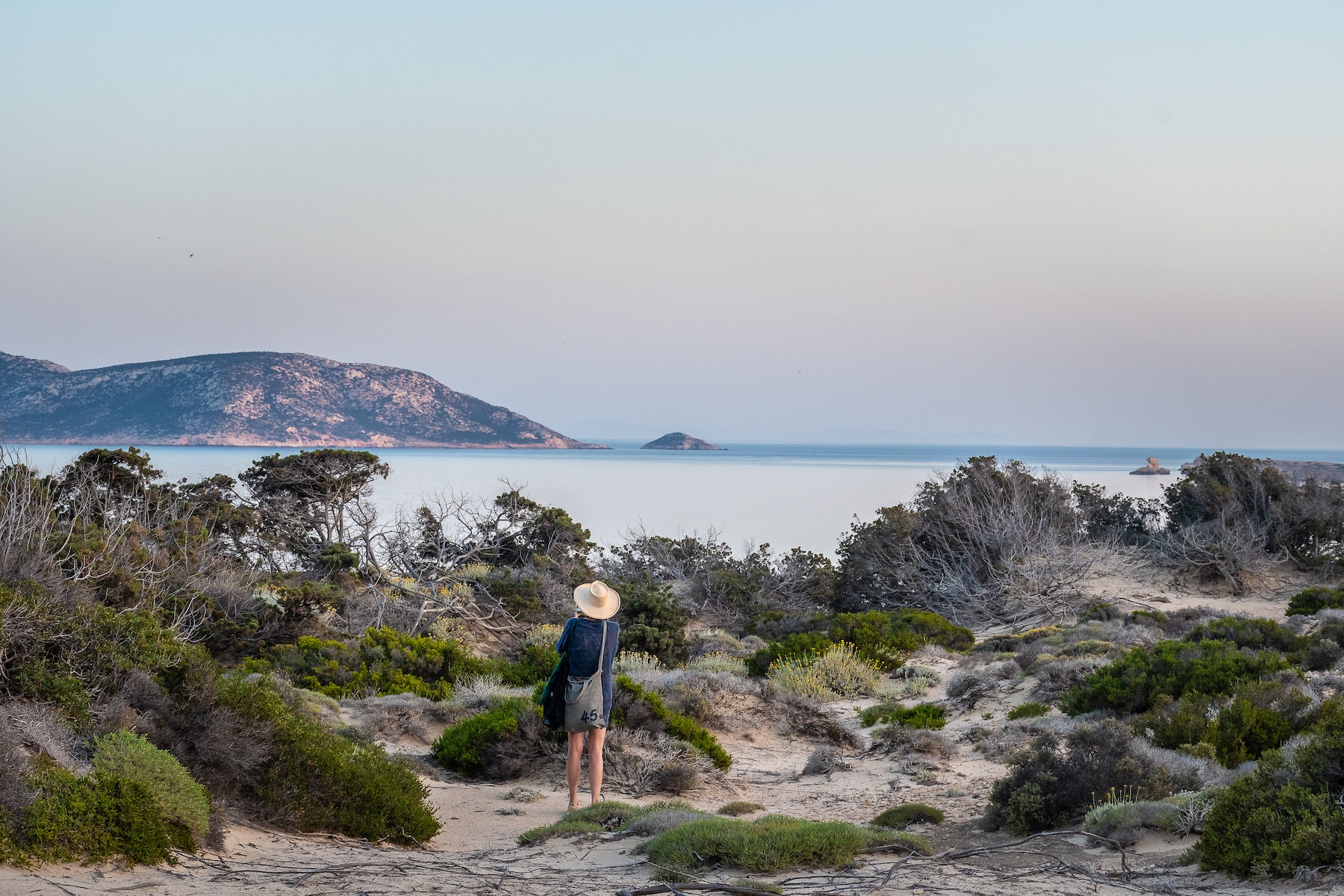 A traveller in a sun hat on the juniper-clad dunes of Koufonisia at dusk, gazing across the calm Aegean to the island of Keros, setting of the boutique villas Éros Kéros.