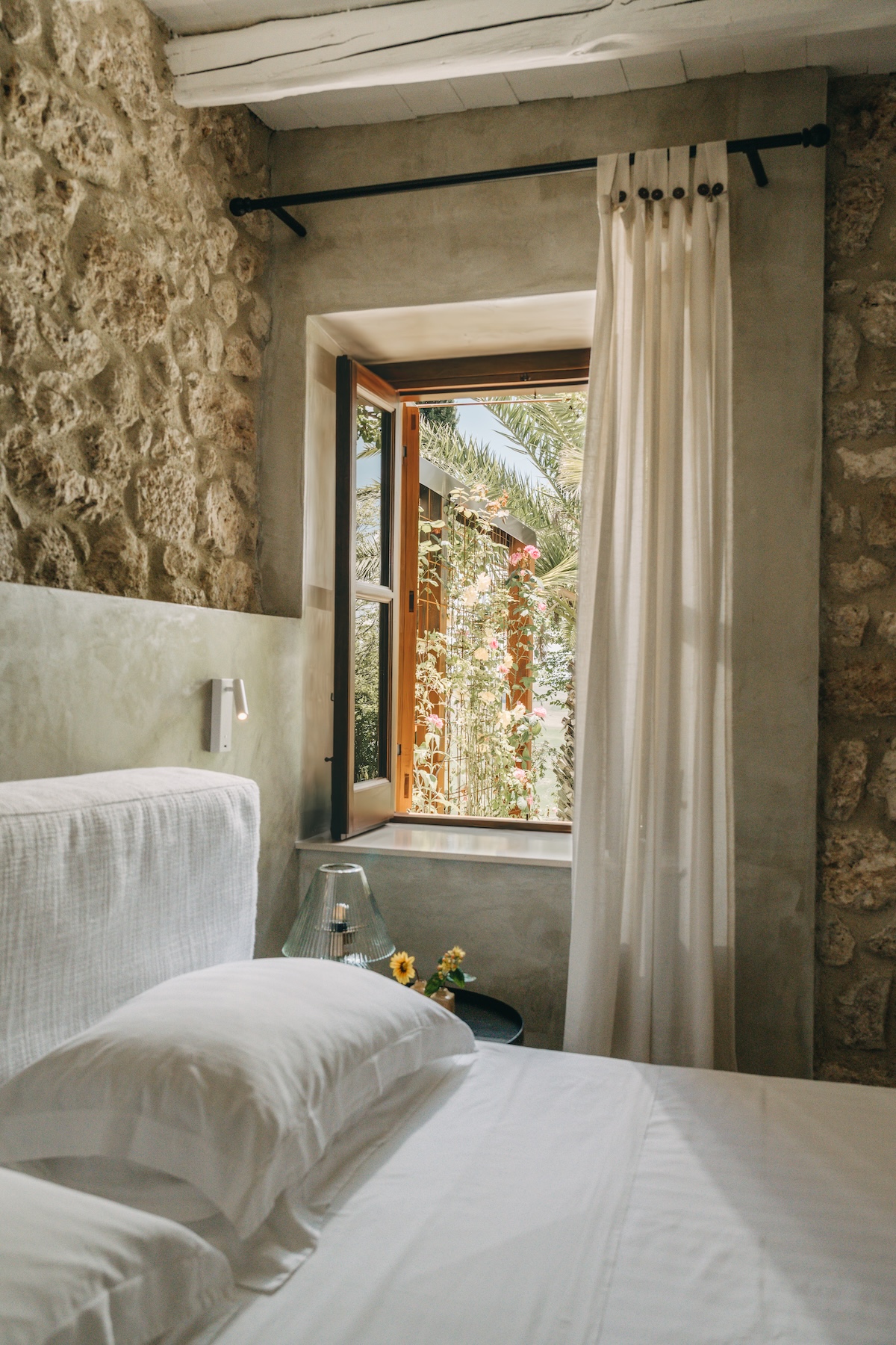 SUSAFA Boutuqie Hotel Sicily |Rough stone bedroom wall and an open window framing climbing roses and palms at a Sicilian masseria