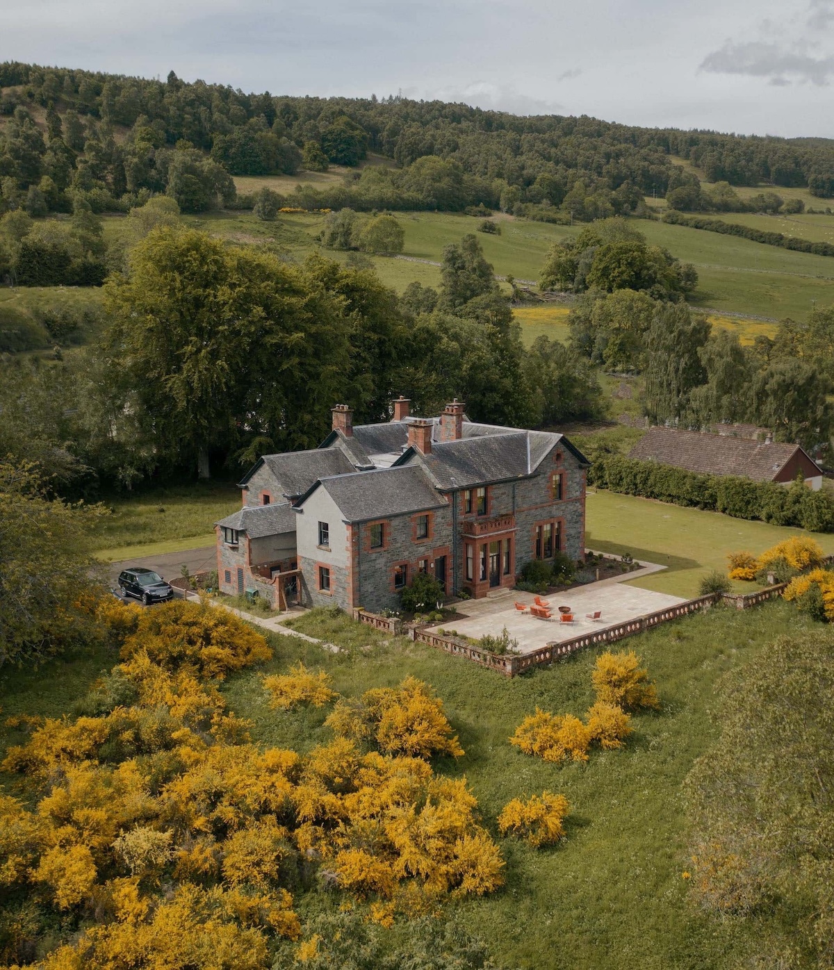Aerial view of Dun Aluinn near Aberfeldy, a Victorian sandstone mansion with a stone terrace and bright orange outdoor furniture, surrounded by gorse and Perthshire meadows.